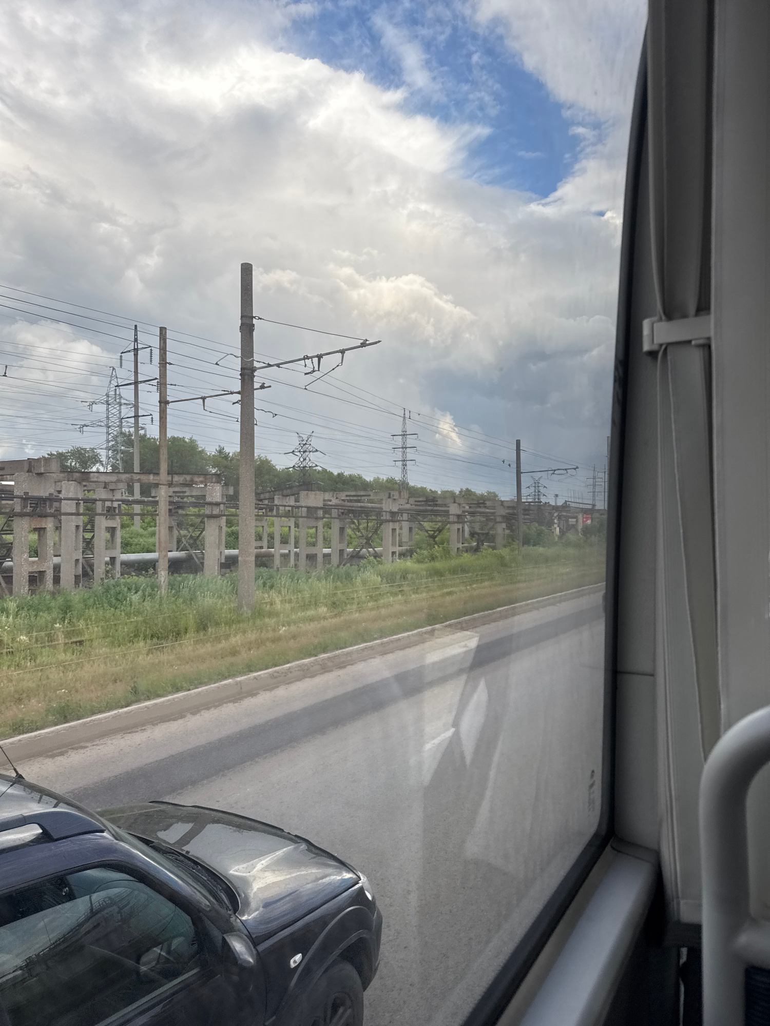 The image shows a view from inside a vehicle, looking out a window. The window appears to be slightly dirty, with smudges and reflections obscuring some details. The scene outside is dominated by a power substation with concrete pylons and overhead wires. The substation extends into the distance, with more pylons and trees visible on either side of a paved road. A dark-colored car is partially visible on the right side of the frame, parked or moving alongside the road.
The sky is cloudy, with patches of blue sky peeking through. The overall lighting suggests it is daytime, likely late morning or early afternoon. The weather appears overcast but not actively raining. The landscape is somewhat industrial and rural, typical of infrastructure along a road in an area like Perm, Russia. There is no discernible text in the image. The image is oriented vertically, with the top of the frame showing the interior of the vehicle, possibly a bus or a large SUV, with a white trim and a handle. The scene outside is viewed from a slightly elevated perspective. No people are visible. The mood is neutral, focusing on the infrastructure and the roadside environment.