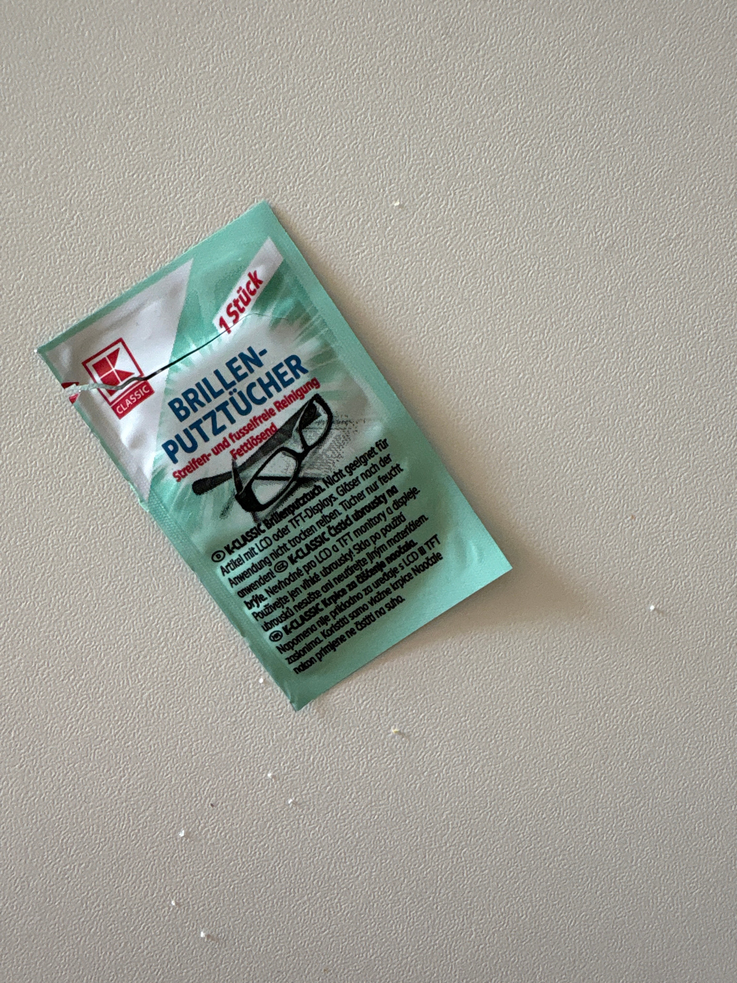 A single, small, teal-colored sachet containing one K-Classic branded eyeglass cleaning wipe is positioned on a light gray, textured surface. The sachet is partially opened, revealing a portion of the wipe inside. The surface appears to be a countertop or table with subtle indentations and a slight sheen. The lighting is even, casting no significant shadows. There are no people, animals, or other objects visible in the frame. The scene is devoid of any contextual clues indicating its location within Bönnigheim, Germany.