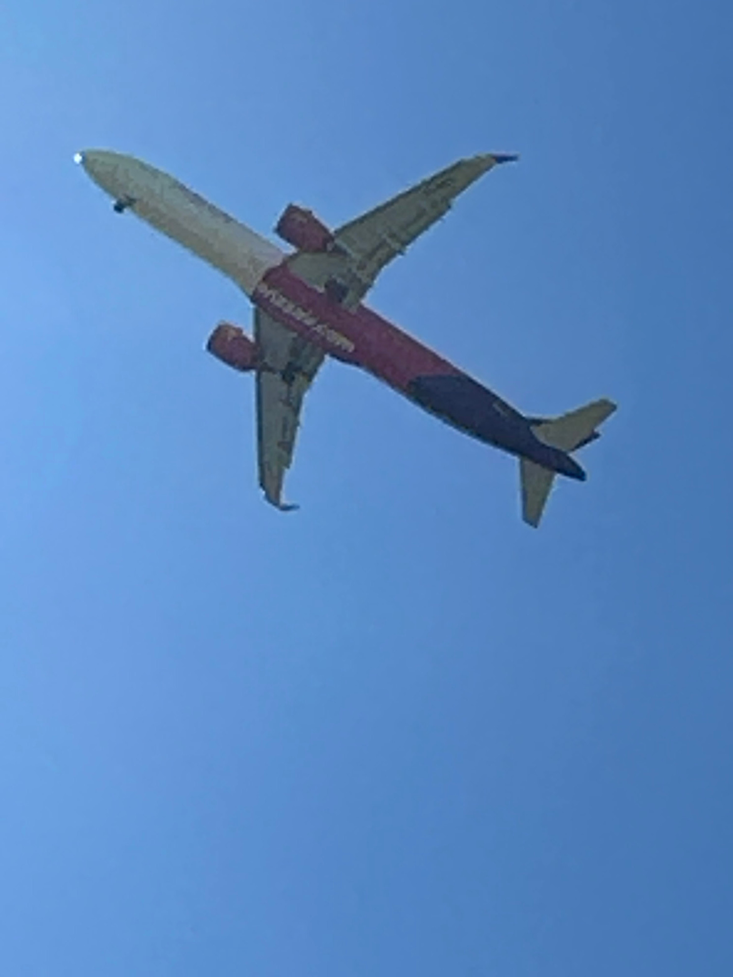 A low-angle, wide shot captures an airplane flying against a clear blue sky. The aircraft is viewed from below, with its wings and tail section prominently visible. The fuselage is primarily colored a light cream, with a dark purple band running along its length and a pink section near the tail. The engines, mounted under the wings, are dark red. The tail fin is also dark purple, with light yellow horizontal stabilizers. A bright white light is visible at the tip of the nose of the airplane. The image appears to be taken from the ground, looking directly upwards. The exact location is unknown.