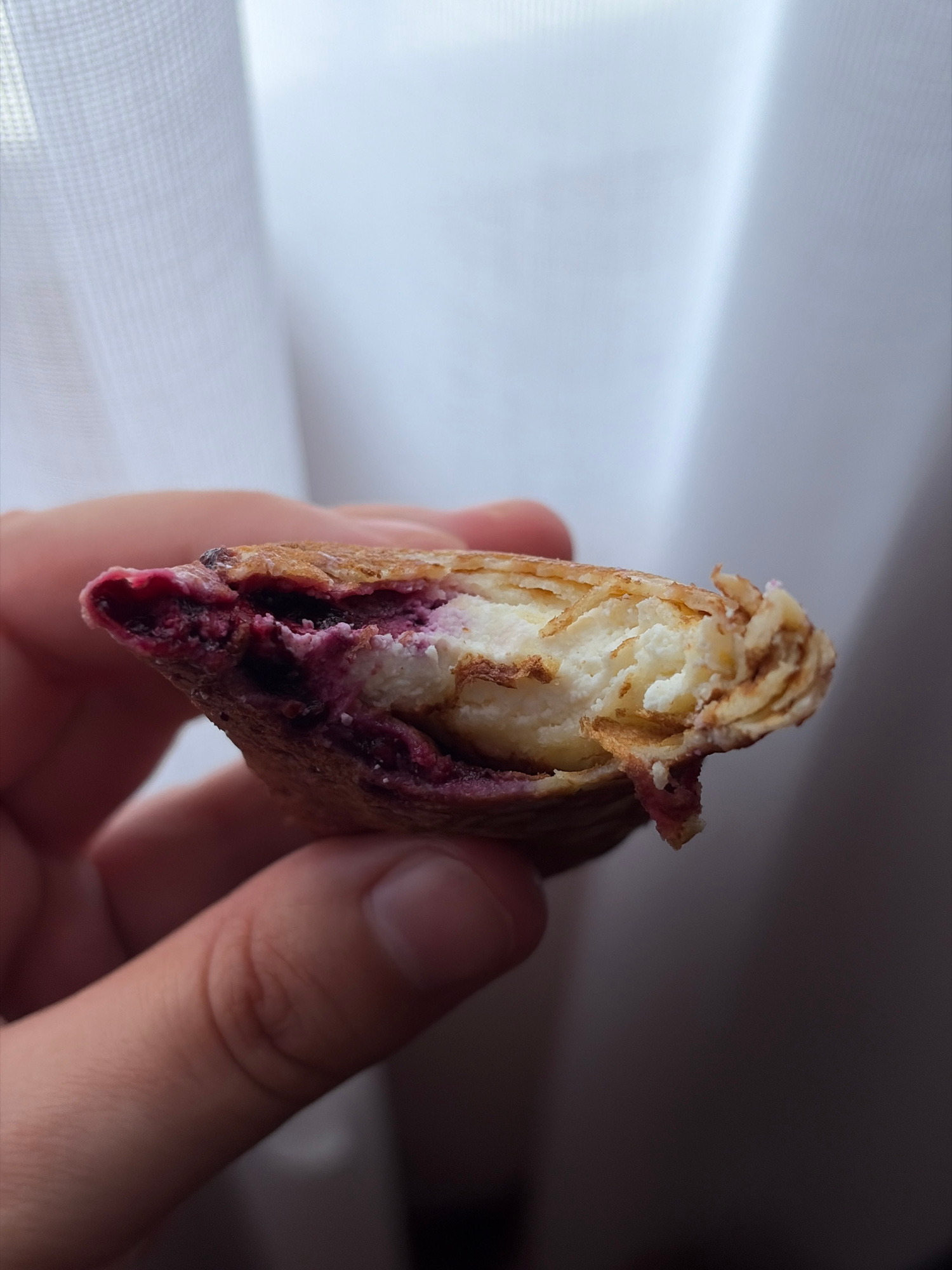 A close-up, side view shows a hand holding a bite-sized pastry filled with cream cheese and blueberries. The pastry is golden brown with flaky layers, and the filling is visible as white cream cheese and deep purple blueberries. The background is blurred white fabric, likely curtains.