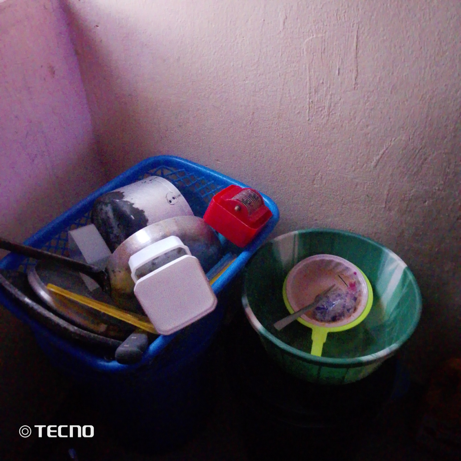 A blue plastic basket is filled with kitchen items. Inside the basket are a large metal pot with a black and silver exterior, a white rectangular container, and a small red object with a rolling wheel. A yellow object, possibly a spatula or serving spoon, is also visible. A portion of a green plastic basin is present to the right of the basket, containing a yellow strainer with a pink rim. A silver utensil is inside the strainer, and a purple substance is visible within the strainer. The objects are situated against a textured, light-colored wall. The bottom left of the image displays a watermark that reads "TECNO". The scene appears to be indoors, likely in a kitchen or storage area, possibly in the city of Jalingo, Nigeria, given the presence of the watermark. There are no people or animals visible in the image.