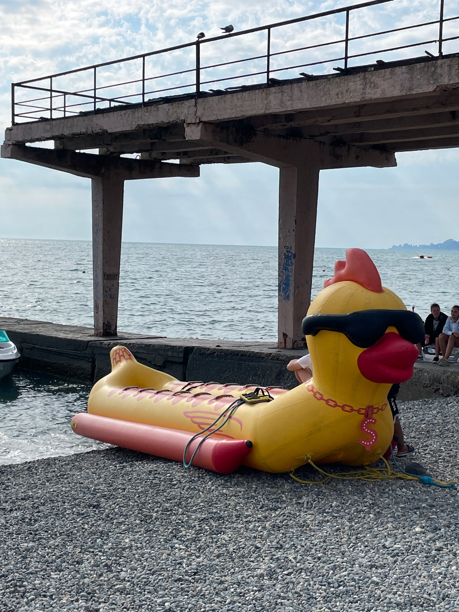 A large, inflatable yellow duck, decorated with pink flame-like patterns and a chain collar, rests on a pebbly shoreline next to the sea. The duck appears to be a novelty item, possibly for water play, as it has a red cap and black sunglasses. Parts of its body are in the water, indicating it's at the edge of the sea.
In the background, several people are visible sitting on a concrete structure that appears to be a pier or a promenade. One person is wearing a white shirt and dark shorts, another a dark jacket and white shirt. They seem to be enjoying the view of the sea. The sea itself is calm, with small ripples on the surface. The sky is overcast with some clouds visible.
To the left of the frame, a dilapidated concrete and metal structure, likely part of an old pier or platform, juts out towards the sea. Its weathered appearance suggests it has been exposed to the elements for a considerable time. The overall scene suggests a casual, possibly tourist-oriented location, given the inflatable toy and the people relaxing by the water. The time of day appears to be daytime, with diffused light characteristic of an overcast sky. The location is identified as Sochi, Russia.