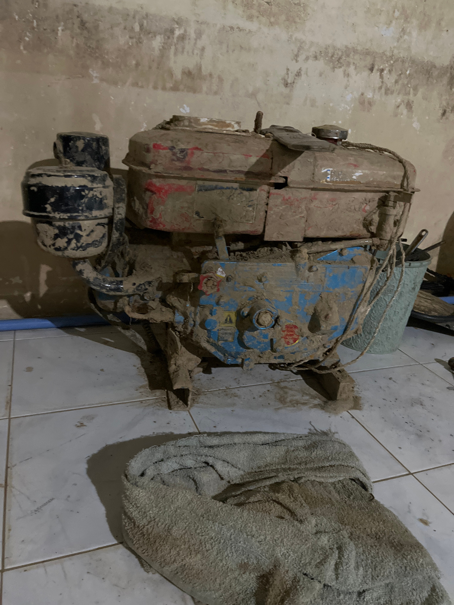 This image shows a dirty, possibly disused, engine. The engine is covered in mud and appears to be an older model, with some red and blue paint visible beneath the grime. It is situated on a tiled floor, with a piece of dirty, grey fabric or towel to its right. The background wall is textured and discolored, suggesting a neglected or damp environment. There are some visible pipes or hoses connected to the engine, one of which is blue. The lighting is dim, and the overall impression is one of disrepair and abandonment. No people are visible in the frame, and no specific activity is taking place. The image is a still photograph, likely taken indoors in a dimly lit space. There is no visible text in the image. Given the location context of Kota Kuala Simpang, Indonesia, the mud suggests a recent flood or prolonged exposure to damp, dirty conditions.
