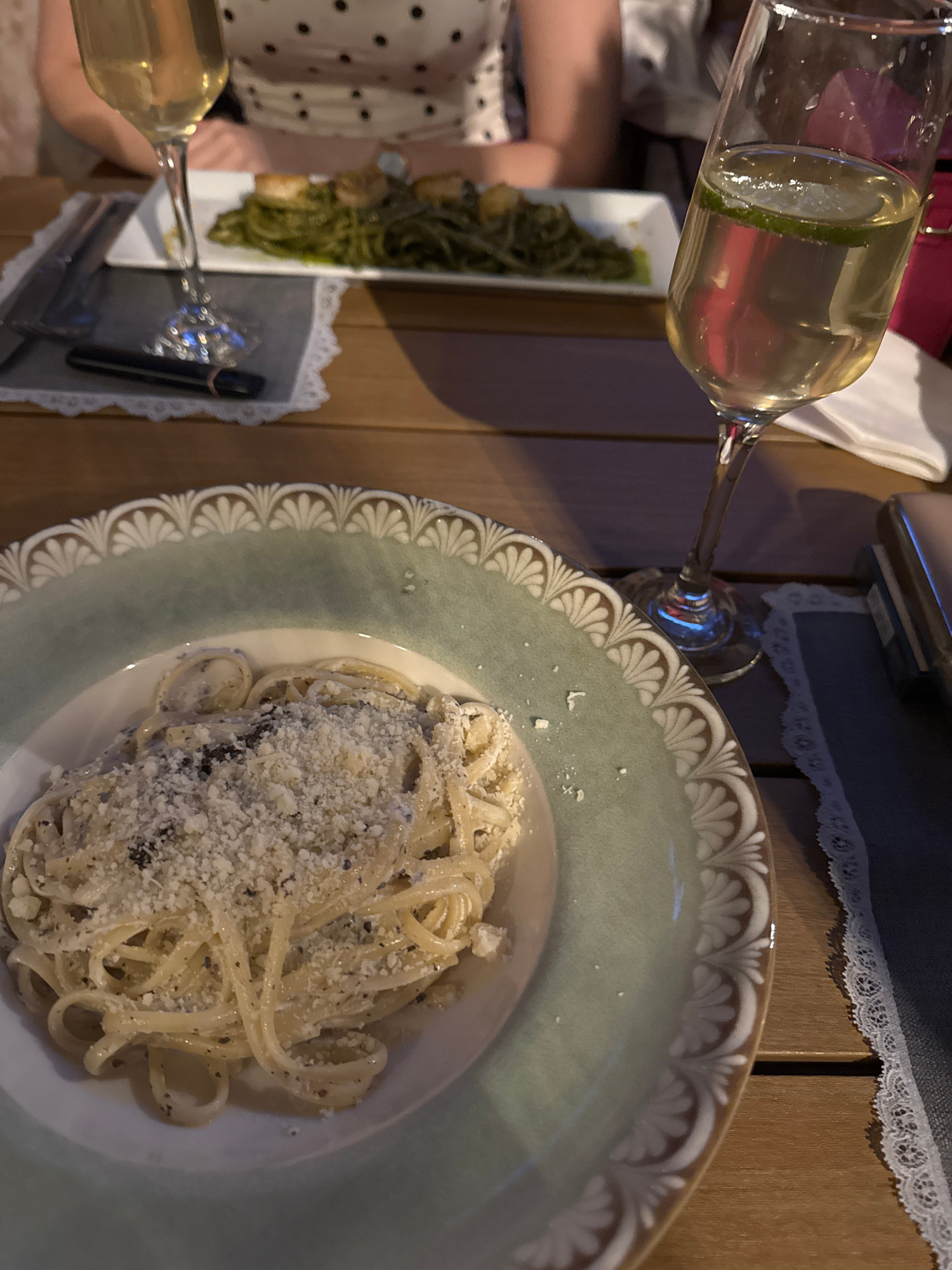 A close-up, high-angle shot captures a table setting in what appears to be a restaurant in Sofia, Bulgaria. In the foreground, a plate of linguine pasta is visible, topped with grated cheese and black pepper. The plate has a decorative rim with a light green and white pattern. To the right of the pasta plate, a champagne flute filled with a pale yellow liquid, garnished with a lime slice, stands on the table. In the background, another plate of pasta, this one dark green with some white toppings, is partially visible along with another champagne flute. A person's torso wearing a black and white polka dot shirt is visible in the upper background. The table is made of wood.