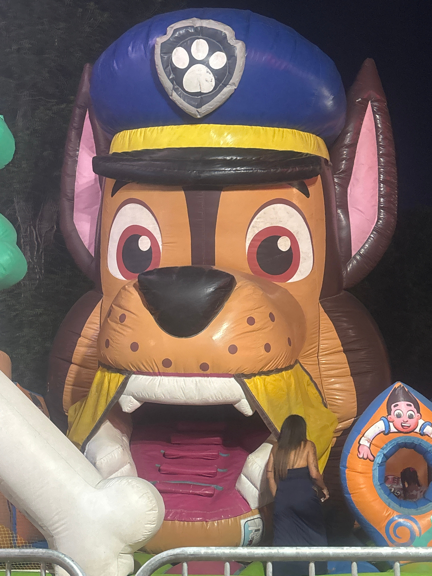 An inflatable structure, designed to resemble the character Chase from the animated show Paw Patrol, is positioned outdoors in the city of Nesebar, Bulgaria. The large inflatable features Chase's signature blue police cap, brown ears, and facial features. Within the inflatable, a slide descends into an opening, and a young girl is visible inside a smaller, orange inflatable tunnel to the right. A woman wearing a dark blue dress stands near the entrance of the main inflatable structure. The background consists of dark foliage and a dark sky, suggesting the image was taken at night. A metal railing is visible in the foreground.