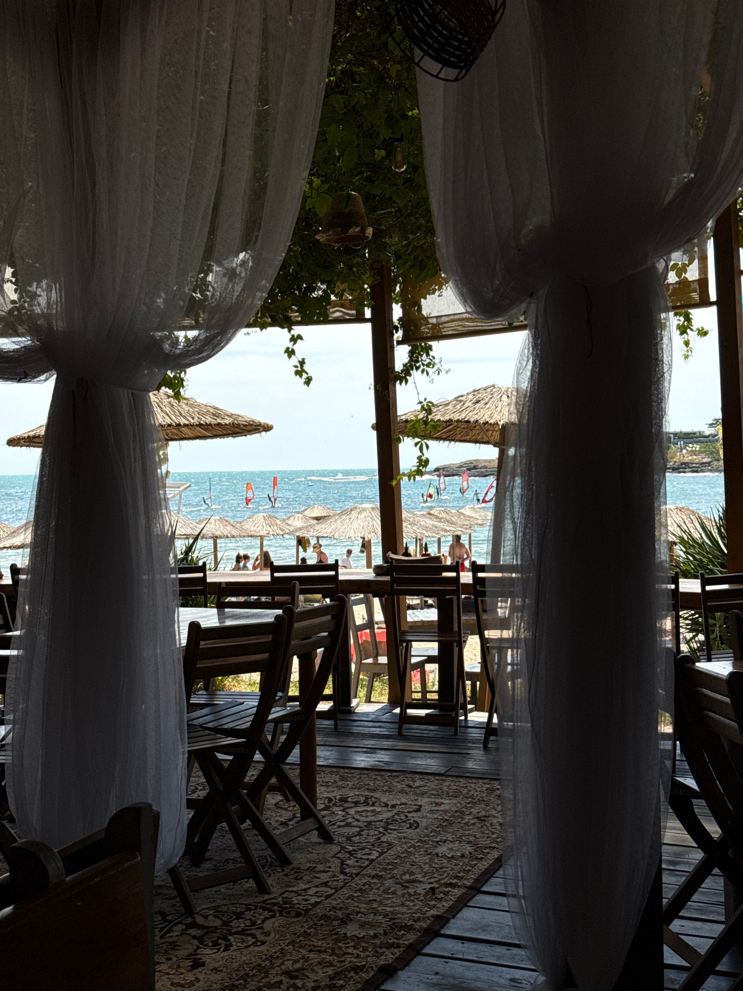 This image captures a view from a covered seating area overlooking a beach. In the foreground, wooden tables and chairs are arranged on a wooden deck covered by an oriental-style rug. Sheer white curtains hang from the ceiling, framing the view. Beyond the seating area, straw-covered umbrellas dot the sandy beach. The sea is visible with several people windsurfing or kitesurfing. The beach is backed by a rocky coastline with some vegetation. The location is identified as the White Lagoon beach in Tsarevo, Bulgaria, a resort known for its water sports.