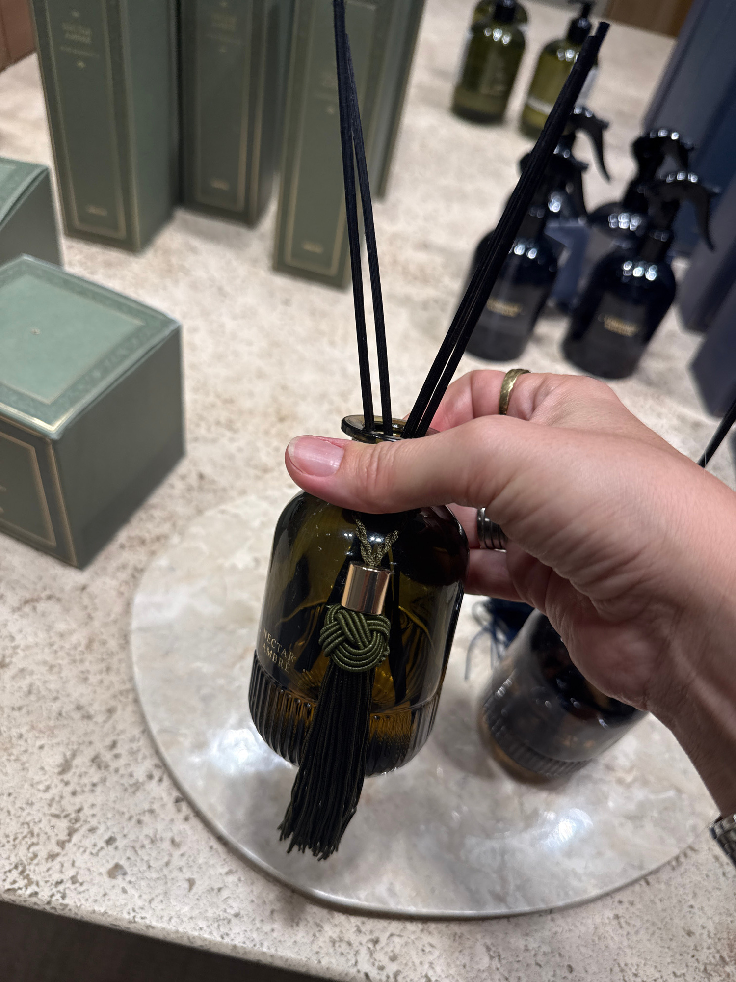 A hand is holding a dark amber glass diffuser with black reeds. The diffuser has a decorative knot and tassel made of dark green string. Text on the diffuser reads "NECTAR AMBRÉ". In the background, there are several other bottles, some with spray tops, and green boxes stacked on a textured surface that appears to be a countertop. The setting seems to be a retail environment, possibly a shop in Almaty, Kazakhstan, displaying luxury home fragrance products. The lighting suggests it could be daytime.