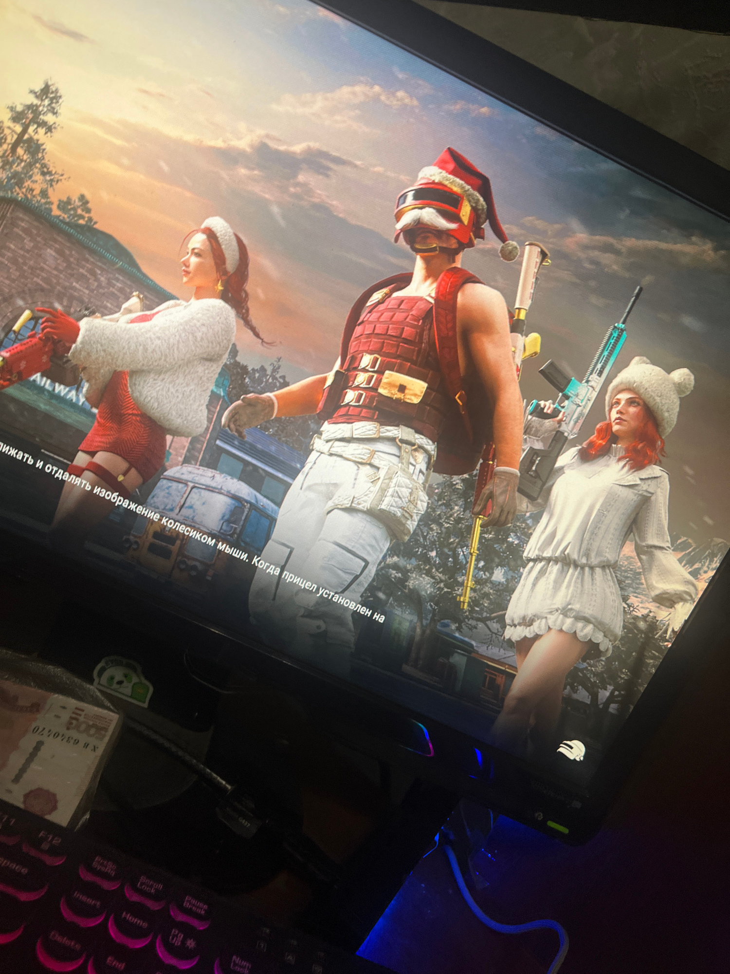 The image displays a computer screen showing a video game with three characters. The characters are posed in front of a blurred background that appears to be a city scene with some trees. The game's interface has some Russian text displayed at the bottom. To the left of the screen, a portion of a keyboard with illuminated keys is visible, along with a small stack of what appears to be cigarette packs. The overall scene is dimly lit, with the primary light source coming from the computer screen and some ambient blue light reflecting from the keyboard. There is no visible indication of the city of Belyye Berega, Russia, within the image itself.