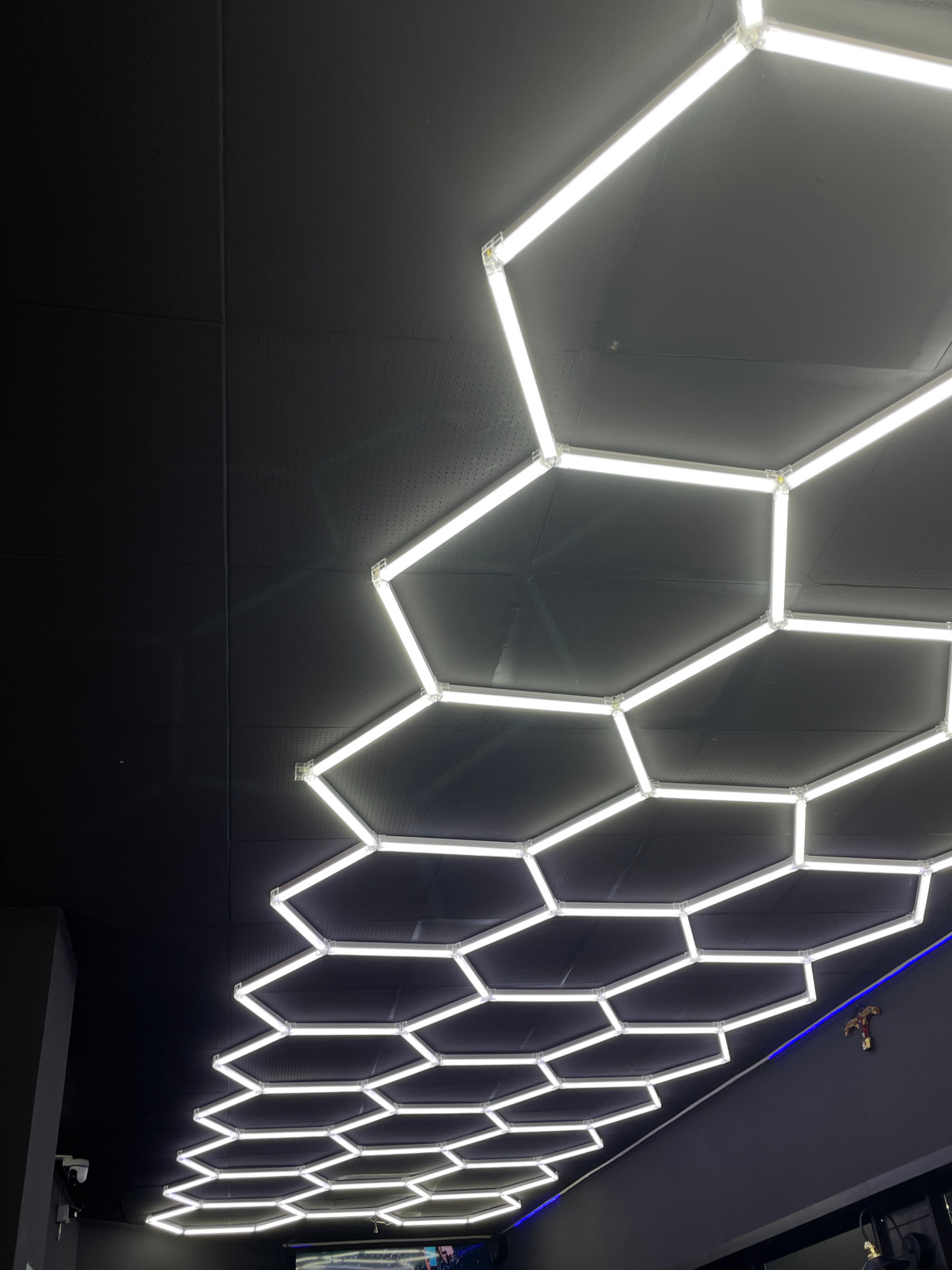 The image shows a wall with a hexagonal light installation. The lights are white and form a repeating pattern of interconnected hexagons. The wall itself is dark gray and appears to be made of large panels. Some panels have a perforated texture, while others are smooth. The lighting from the hexagons casts a glow on the wall. In the upper right corner of the image, there is a glimpse of a blue light strip running along a ceiling or upper wall. There is also a small, brown, figurine-like object perched on the blue light strip. The lower portion of the image shows a dark gray surface, possibly a counter or desk, with a white device on it, which could be a scanner or a payment terminal. The overall setting appears to be indoors, possibly a retail space, exhibition, or a modern office. There is no visible text in the image. The time of day and weather are not discernible. The scene is lit by the artificial light from the installation.