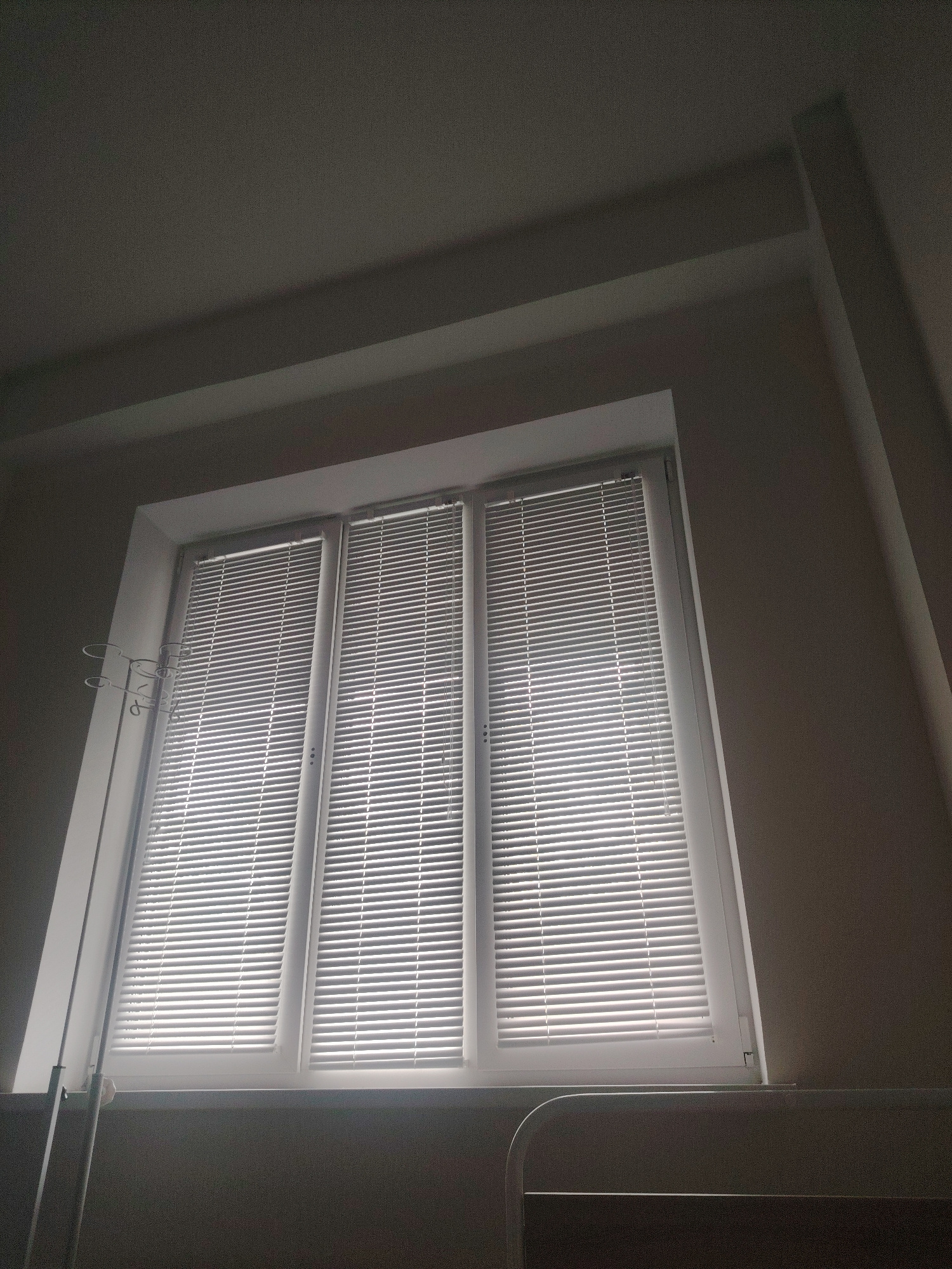 An indoor, eye-level shot captures a three-panel window with white horizontal blinds. The window is framed by a white border and is illuminated by sunlight filtering through the blinds. To the left of the window, a metal frame with multiple circular hooks is attached to the window frame. The room appears to be a hospital room, with a bed frame visible in the foreground. The ceiling is angled and has recessed lighting. The scene is located in a hospital in Mahilyow, Belarus.