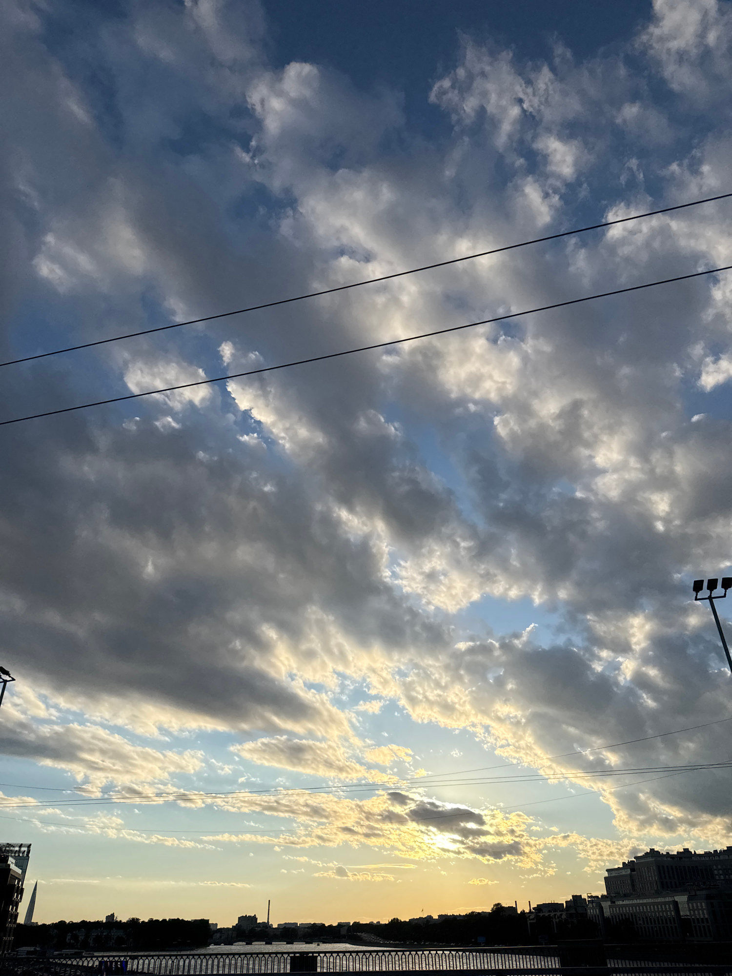 The image captures a dramatic sky filled with dynamic, textured clouds, illuminated by the warm glow of the setting sun. The clouds range in color from deep grays and blues to bright whites and yellows, creating a sense of depth and movement. Faint, dark lines, likely power lines, traverse the sky horizontally.
In the lower portion of the image, a silhouetted cityscape is visible along a body of water. A railing or fence runs along the foreground, suggesting a vantage point near the water's edge. Buildings of various sizes and architectural styles dot the skyline, some appearing quite tall. The sunlight casts a soft, hazy glow over the entire scene, with the sun itself partially obscured by clouds near the horizon. A tall, slender spire-like structure is also discernible on the left side of the cityscape. The overall impression is one of a city at dusk, with the natural beauty of the sky dominating the view.