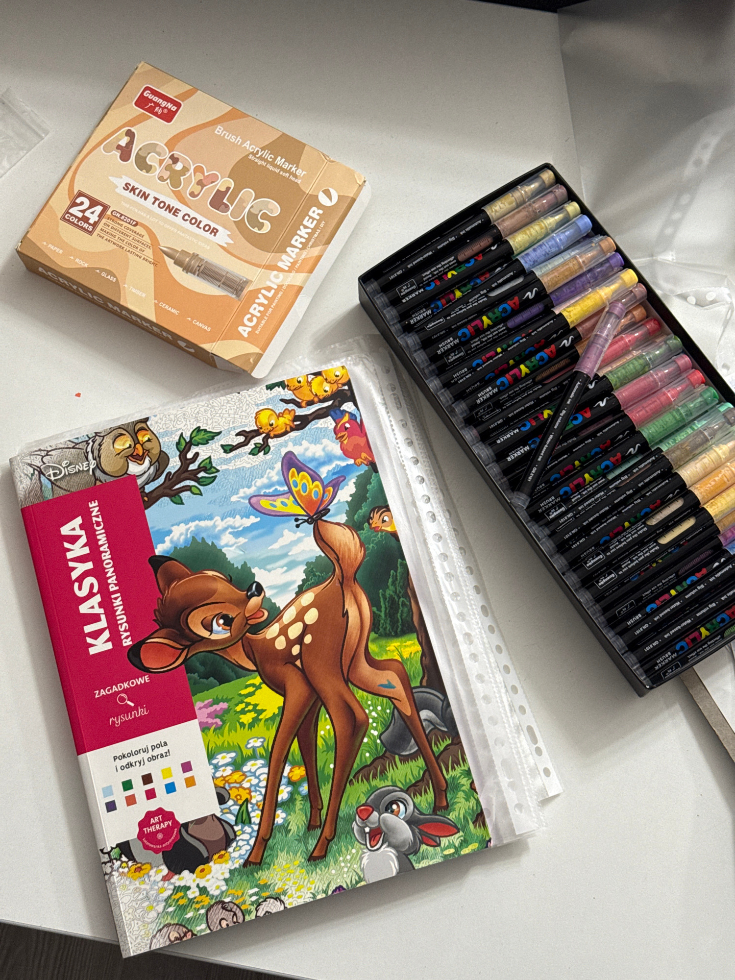 The image displays a white, flat surface, likely a table, with several art supplies. In the upper left corner is a box labeled "GuangNa ACRYLIC BRUSH Acrylic Marker SKIN TONE COLOR 24 COLORS". Next to it, on the right, is a black plastic case containing a set of multi-colored acrylic markers. In the center and lower portion of the image is a children's coloring book featuring characters from Disney's Bambi, including Bambi, Thumper, and various forest animals. The coloring book has a pink cover with Polish text that reads "KLASYKA RYSUNKI PANORAMICZNE" and "ZAGADKOWE rysunki". The overall scene suggests a creative activity or hobby. There is no discernible information to indicate a specific location in the city of Łódź, Poland.