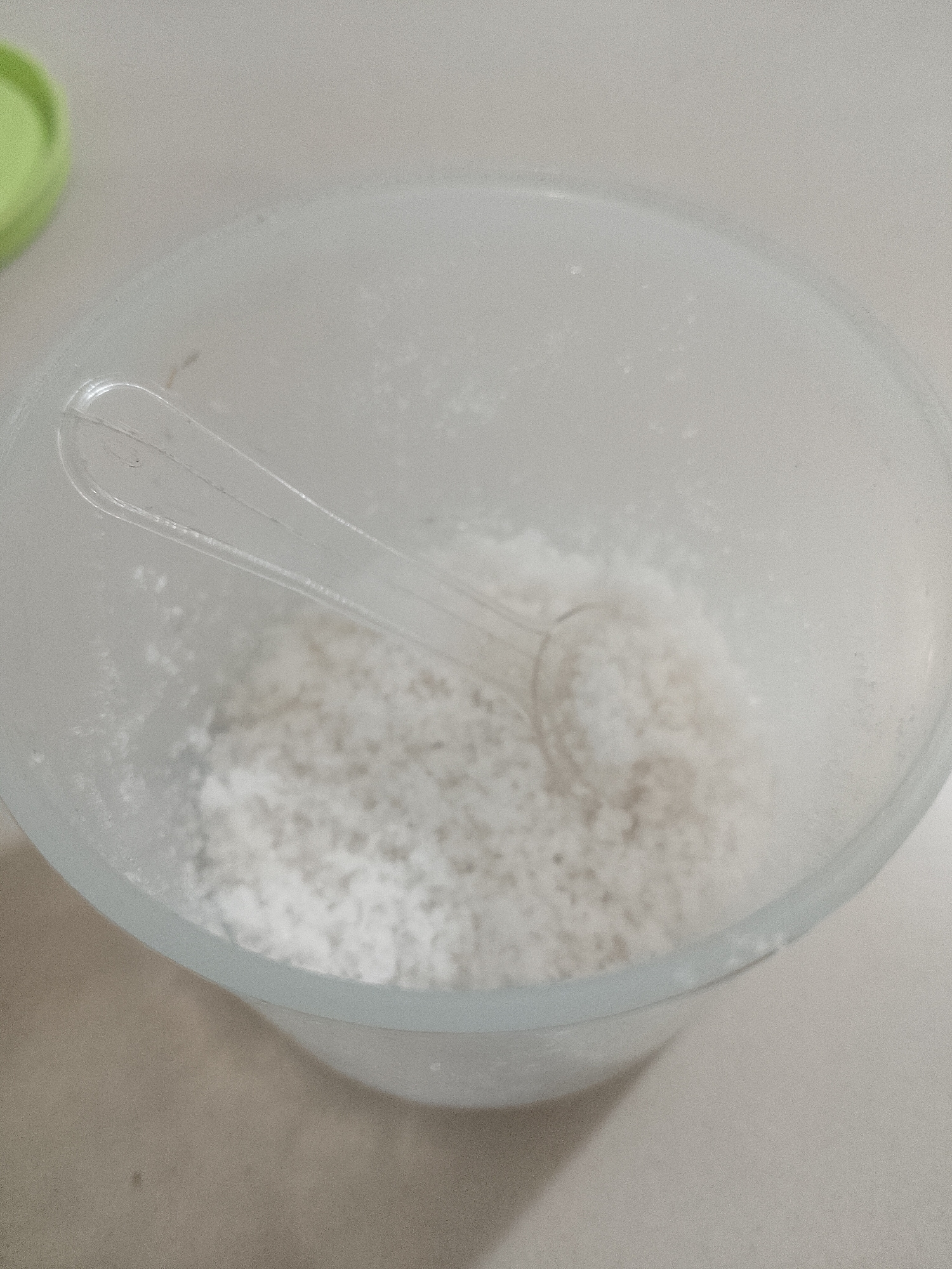 A clear plastic container filled with coarse white material, likely salt, sits on a light-colored surface. A clear plastic scoop with a slightly curved handle rests inside the container, partially submerged in the material. The container has a frosted appearance. In the upper left corner of the image, a portion of a lime green lid is visible. The background is a neutral, light-colored surface. The image is taken from a top-down perspective, focusing on the contents of the container. There is no discernible location in Lhokseumawe, Indonesia.