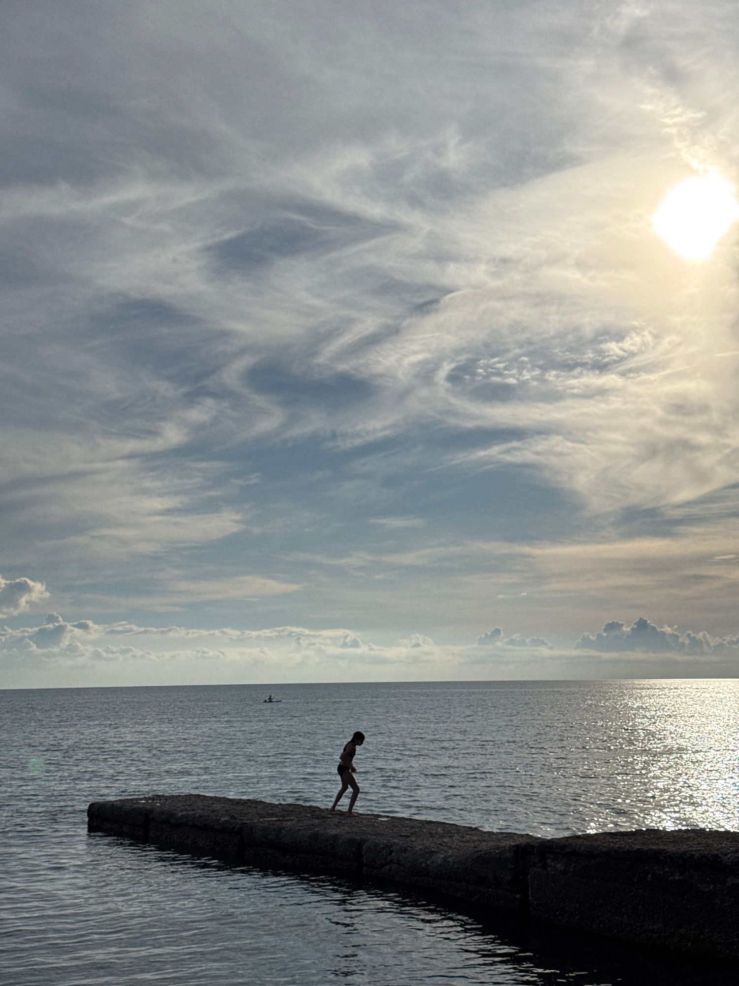 A solitary child in silhouette stands on a concrete pier extending into a calm, dark sea. The child is facing away from the viewer and appears to be walking or preparing to jump into the water. The sky above is dramatic, filled with wispy, white clouds that are illuminated by the bright, diffused light of the sun. The sun itself is partially obscured by clouds, creating a lens flare effect. In the distance, two figures on a small boat or kayak are visible on the water, adding a sense of scale. The scene suggests a serene, late afternoon or early evening ambiance, possibly at one of Sochi's many piers along the Black Sea coast. The water reflects the shimmering sunlight, creating a textured surface.