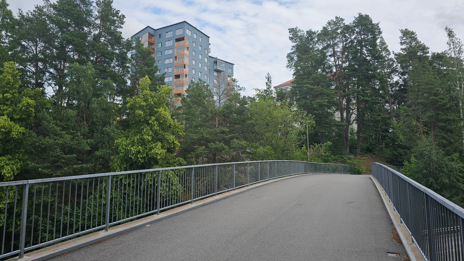 The image shows a pedestrian bridge with a grey asphalt surface and a metal railing. The bridge curves to the right, leading into a densely forested area. Behind the trees, several apartment buildings with light blue facades and orange balconies are visible. The sky is cloudy. The location is likely an urban area in Linköping, Sweden, given the presence of apartment buildings and the characteristic pine trees.