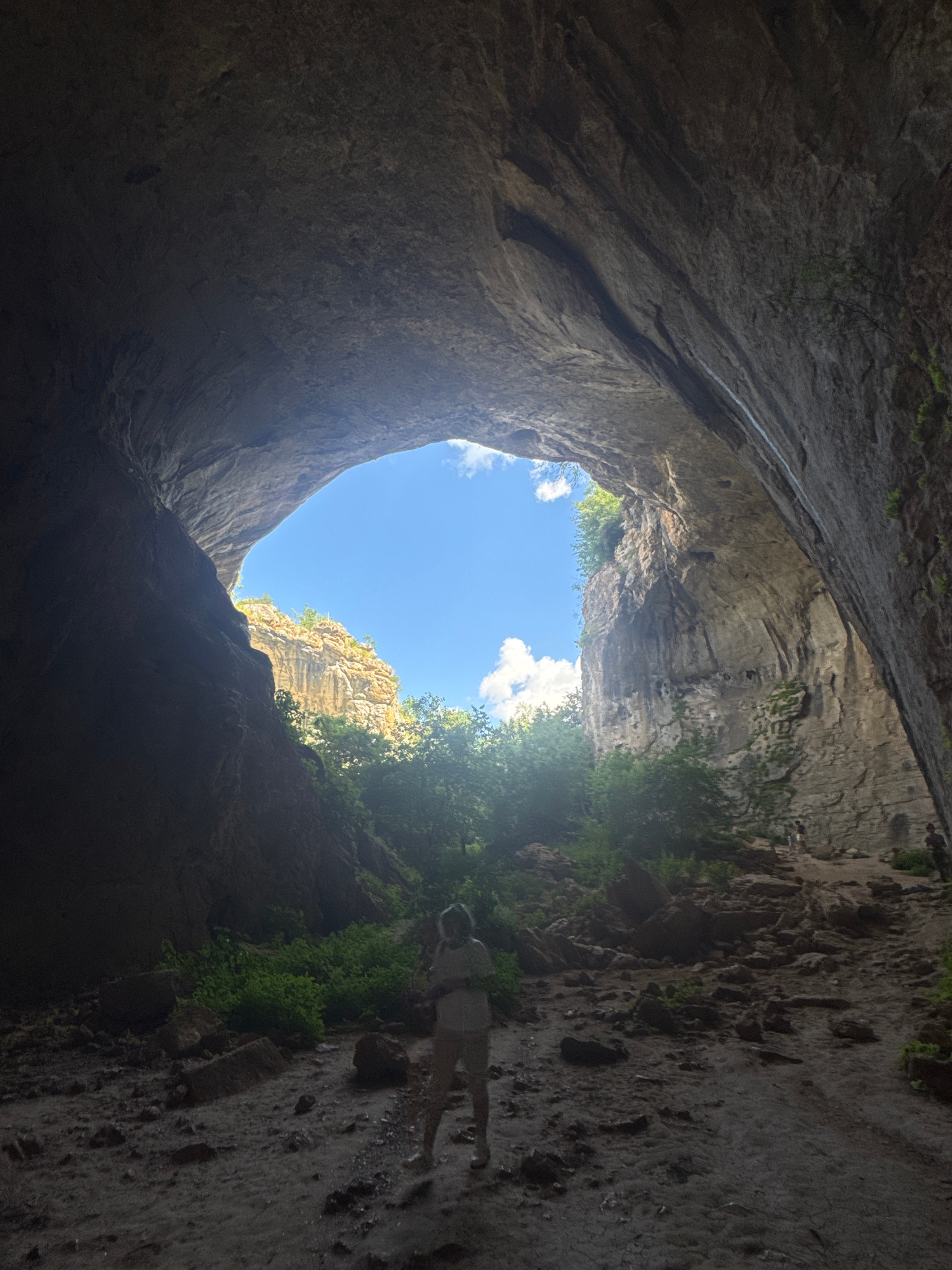 The image captures a view from within a large cave opening, looking out towards a bright blue sky with scattered white clouds. The cave ceiling arches overhead, revealing textured rock formations. Sunlight streams in, illuminating the lush green foliage and rugged rock faces outside the cave. In the foreground, a person stands with their back to the camera, facing the exit of the cave. The ground is covered in rocks and dirt. The overall scene suggests an exploration of a natural wonder, likely a cave in the Karlukovo region of Bulgaria, known for its karst formations.