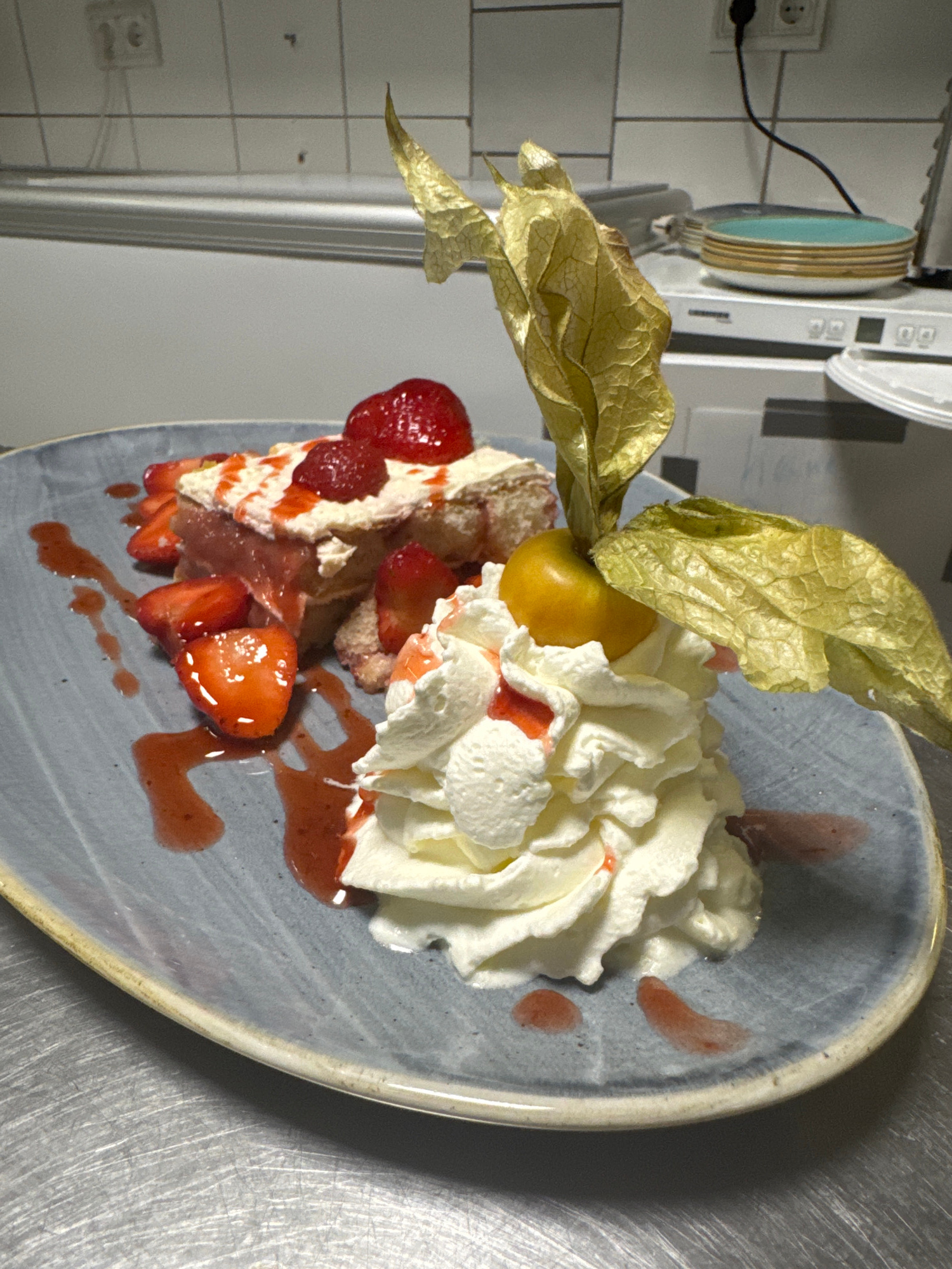 A close-up, slightly elevated shot captures a dessert presentation on a blue-grey textured plate. The dessert consists of a layered cake or pastry topped with whipped cream and fresh strawberries. Sliced strawberries and a strawberry sauce are artfully arranged around the plate, creating a vibrant red contrast. A single physalis fruit, with its characteristic papery husk, is perched atop the whipped cream, serving as a garnish. The background, out of focus, suggests a commercial kitchen environment with stainless steel surfaces and tiled walls, including electrical outlets. The overall impression is of a carefully prepared and aesthetically pleasing dessert.