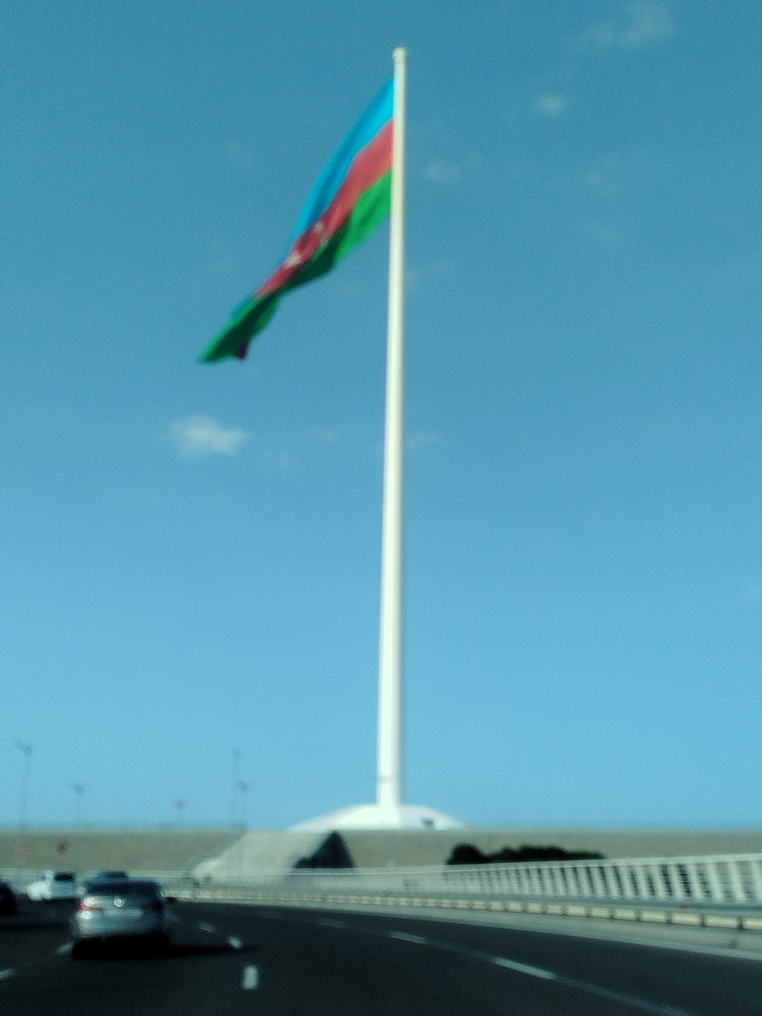The image captures a view of the Azerbaijan National Flag hoisted on a tall flagpole, situated in the city of Baku, Azerbaijan. The flag, composed of three horizontal bands of blue, red, and green, with a white crescent and eight-pointed star on the red band, is prominently displayed against a clear blue sky. The flagpole stands on a large, white circular base. In the foreground, a highway is visible with several cars driving on it. The overall scene suggests a modern urban environment with a significant landmark.
