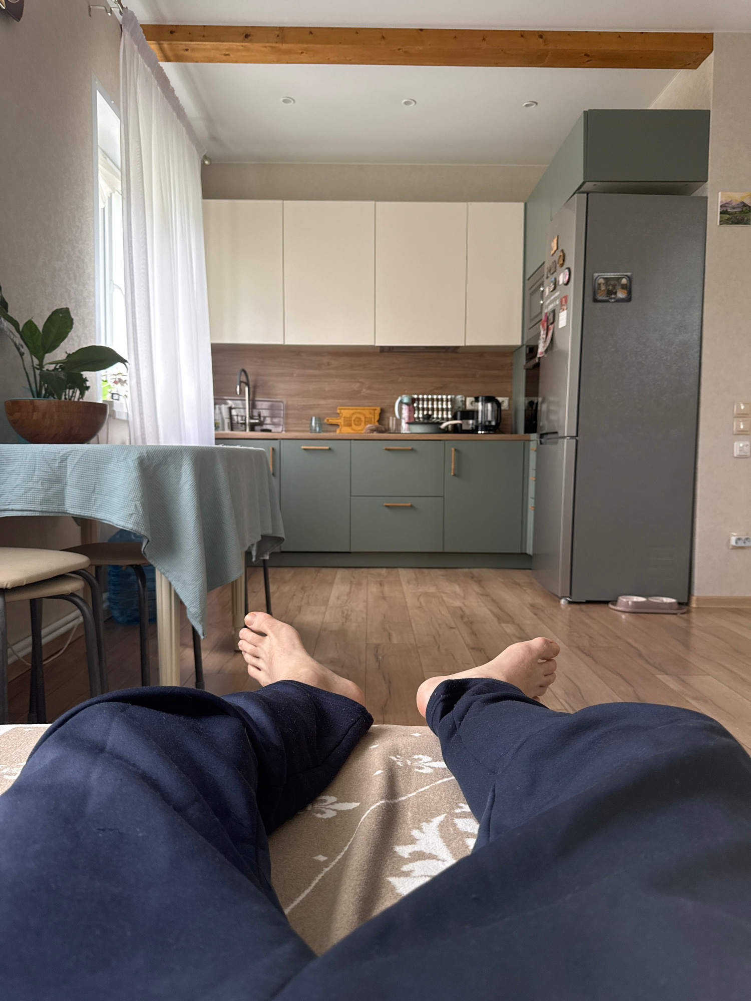 This is a first-person perspective image taken from a reclining position, looking towards a kitchen area. The foreground shows a person's navy blue pants and bare feet resting on a beige blanket with a floral pattern. The middle ground features a dining table covered with a light blue tablecloth, partially obscuring two stools. A large houseplant in a wooden pot is visible on the table. Beyond the table, a modern kitchen is depicted with light beige cabinets on the top and sage green cabinets on the bottom. A wooden backsplash runs along the wall behind the countertops. Appliances like a kettle and a coffee maker are on the counter, and a stainless steel refrigerator stands to the right. A window with sheer white curtains is visible on the left side of the frame. The flooring throughout the visible area is light-colored wood. The scene appears to be an interior residential setting, potentially a kitchen and dining area within a home in Bun'kovo, Russia.