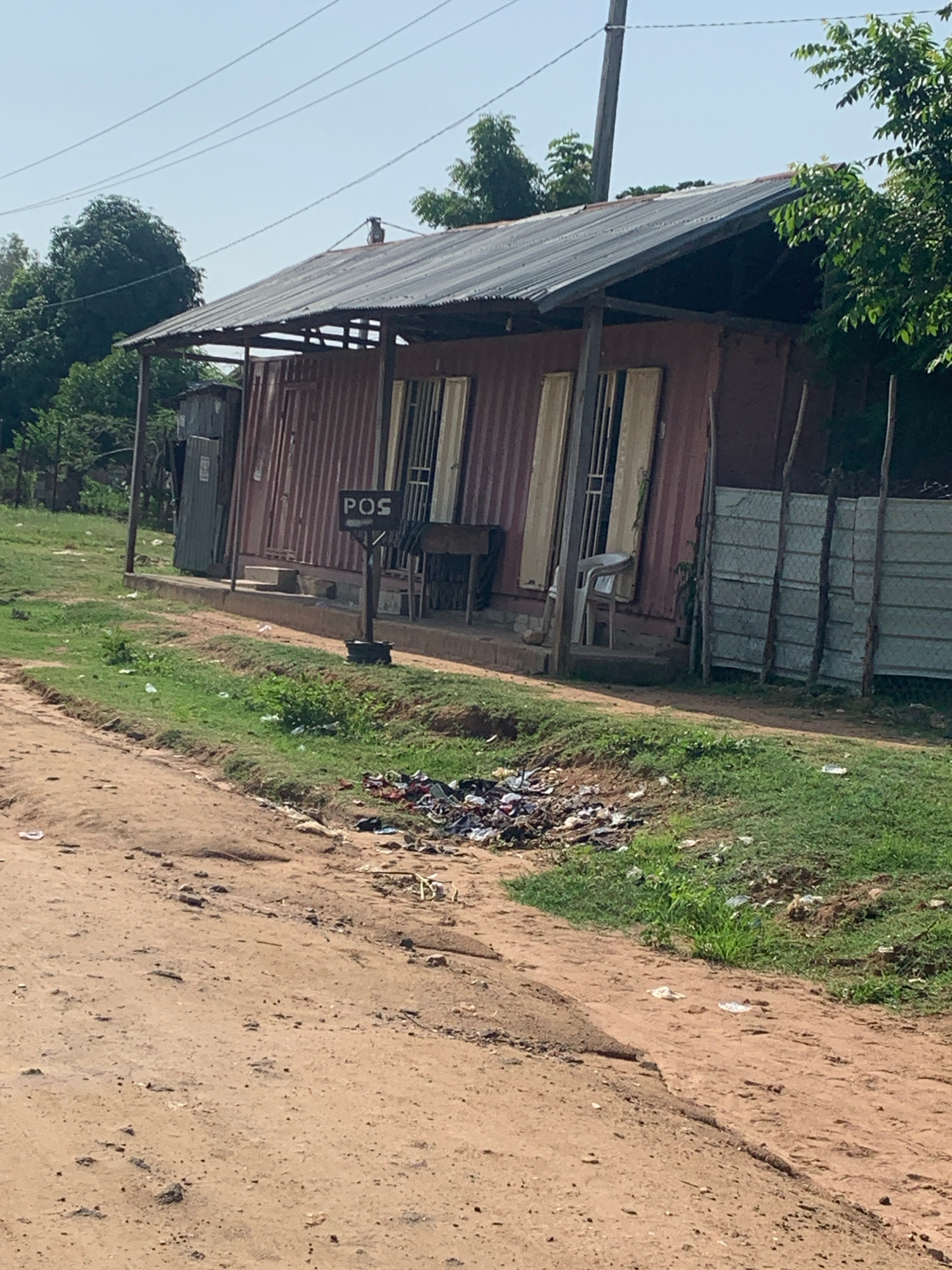 This image depicts an outdoor scene, likely in a rural or developing area, under bright daylight conditions. The primary subject is a small, rustic structure, possibly a shop or kiosk, constructed from corrugated metal and what appear to be repurposed shipping containers. The building has a sloped, dark-colored roof that extends over an open-air section, supported by metal poles. Various windows and doors are visible on the front of the structure. A white plastic chair is positioned outside.
Next to the building, a pile of refuse and litter is scattered on the ground, suggesting a lack of proper waste disposal in the immediate vicinity. The ground is a mix of dirt and sparse green grass. A vertical sign with the letters "POS" is mounted on a pole in front of the building, indicating a point of sale.
In the background, more trees and vegetation are present, and power lines are visible stretching across the sky, which is clear and blue. The overall impression is one of a functional but somewhat unkempt commercial space in an environment that is not heavily urbanized. No people are visible in the image, and the scene appears static.