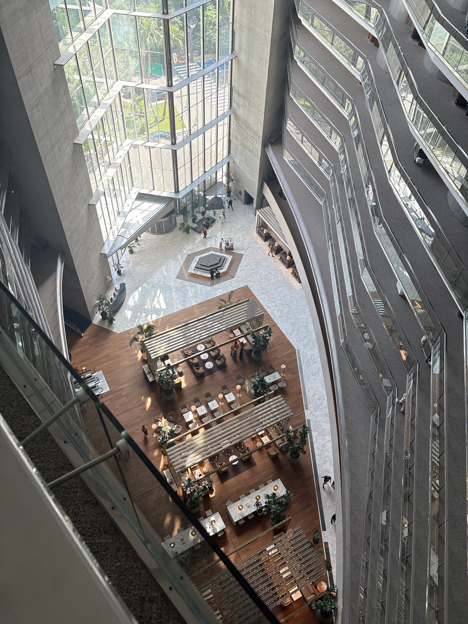 This is a high-angle, full shot looking down into a large, multi-story interior atrium space. The atrium features a massive, angled concrete wall on the left and a series of stepped balconies with glass railings on the right, overlooking the central open space. The atrium is filled with natural light streaming in from a large, polygonal glass-paneled ceiling and wall on the far side.
The central area of the atrium is a large open space with polished marble floors and wooden flooring. On the marble floor, there is a distinctive octagonal water feature or seating area with people gathered around it. Further into the atrium, on the wooden floor, there are several seating areas with tables and chairs, reminiscent of a hotel lobby or restaurant. Some of these areas are shaded by slatted wooden structures. Lush green plants are strategically placed throughout these seating areas, adding a touch of nature.
The stepped balconies on the right are covered with grey carpet and feature glass railings. Several people are visible on these balconies, walking and standing. The perspective suggests these are pathways or seating areas on different levels of the building.
On the far side, through the large glass windows, a glimpse of the exterior is visible, showing greenery and trees, suggesting an urban setting with some landscaping. This could be the Hotel Arts in Barcelona, Spain, known for its striking atrium design and glass facade.
In the foreground, a portion of a glass railing and the edge of a floor from a higher level is visible, indicating the vantage point from which the photo was taken.
There are approximately 10-15 individuals visible throughout the scene, mostly in small groups or walking individually. They are dressed in casual attire. The overall impression is one of a modern, airy, and sophisticated indoor public space.