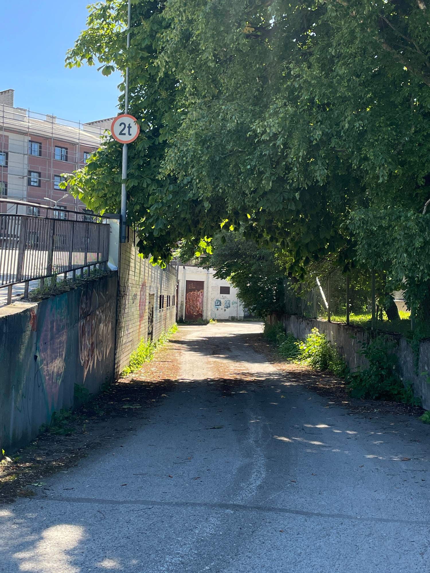 The image captures a narrow, paved pathway bordered by a brick wall covered in graffiti on the left and lush green trees overhead. The wall is interspersed with small, dark openings. Sunlight casts dappled shadows on the pathway, suggesting it is daytime. A sign with "2t" in a red circle is visible amidst the leaves, indicating a weight limit. In the background, a building with scaffolding and windows can be seen, and further down the path, a weathered industrial-looking building with a large, dark door is present. The scene appears to be in an urban or semi-industrial area, possibly an alleyway or a service road, with a clear sky suggesting fair weather. No people are visible in the frame, and the overall atmosphere is quiet and somewhat desolate. The graffiti on the wall adds a touch of urban decay. The location is identified as Tallinn, Estonia.