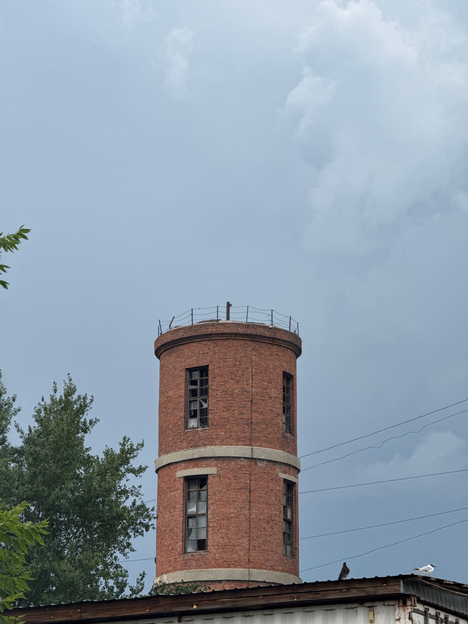 An old, circular brick tower with broken windows stands against a cloudy sky. A few green trees are visible to the left, and power lines stretch across the upper portion of the frame. The tower appears to be part of an abandoned industrial complex. There are two birds perched on the roof of a nearby structure in the lower right corner of the image.