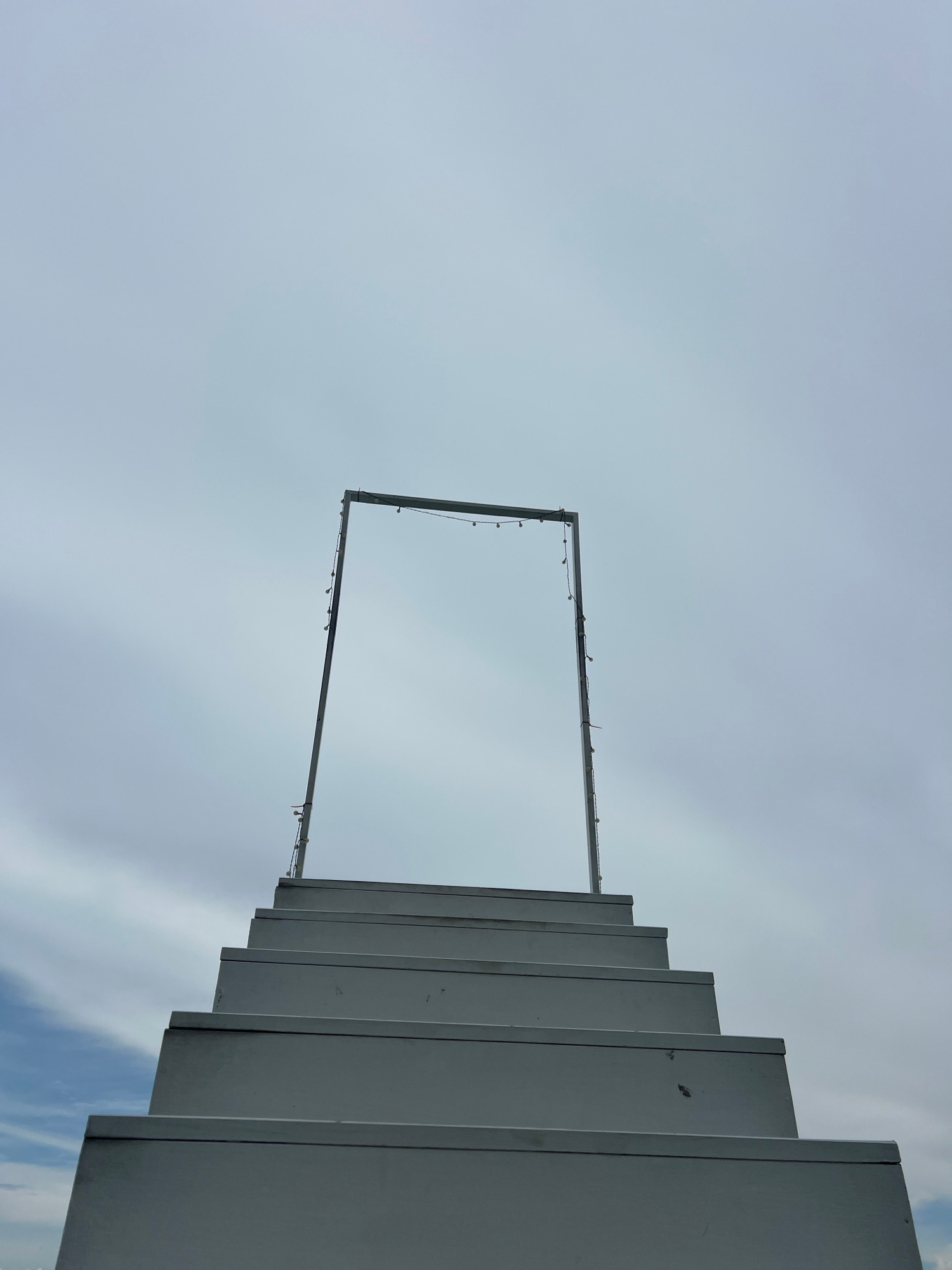 The image shows a minimalist, abstract composition set against a cloudy sky. The primary subjects are architectural elements and a string of lights. On the right side of the frame, a series of parallel, light-colored, rectangular structures are arranged in a stepped pattern, suggesting a modern staircase or tiered platform. These structures are smooth and appear to be made of a uniform material, possibly metal or painted concrete.
Dominating the upper portion of the image is a thin, rectangular frame made of metal. Along the bottom edge of this frame, a string of small, white lights is draped, with some bulbs visible. The frame is positioned centrally in the sky, creating a sense of an opening or a window.
The sky is overcast with light gray clouds, with a hint of blue sky and wisps of white clouds visible at the bottom right, indicating daytime. The overall mood is calm and serene, with a focus on geometric forms and the interplay of light and shadow. The orientation of the image is vertical, with the architectural elements leading the eye upwards. There are no people visible in the image, and no discernible text. The setting appears to be an outdoor space, possibly an art installation or part of a modern building in Baan Thurian, Thailand.