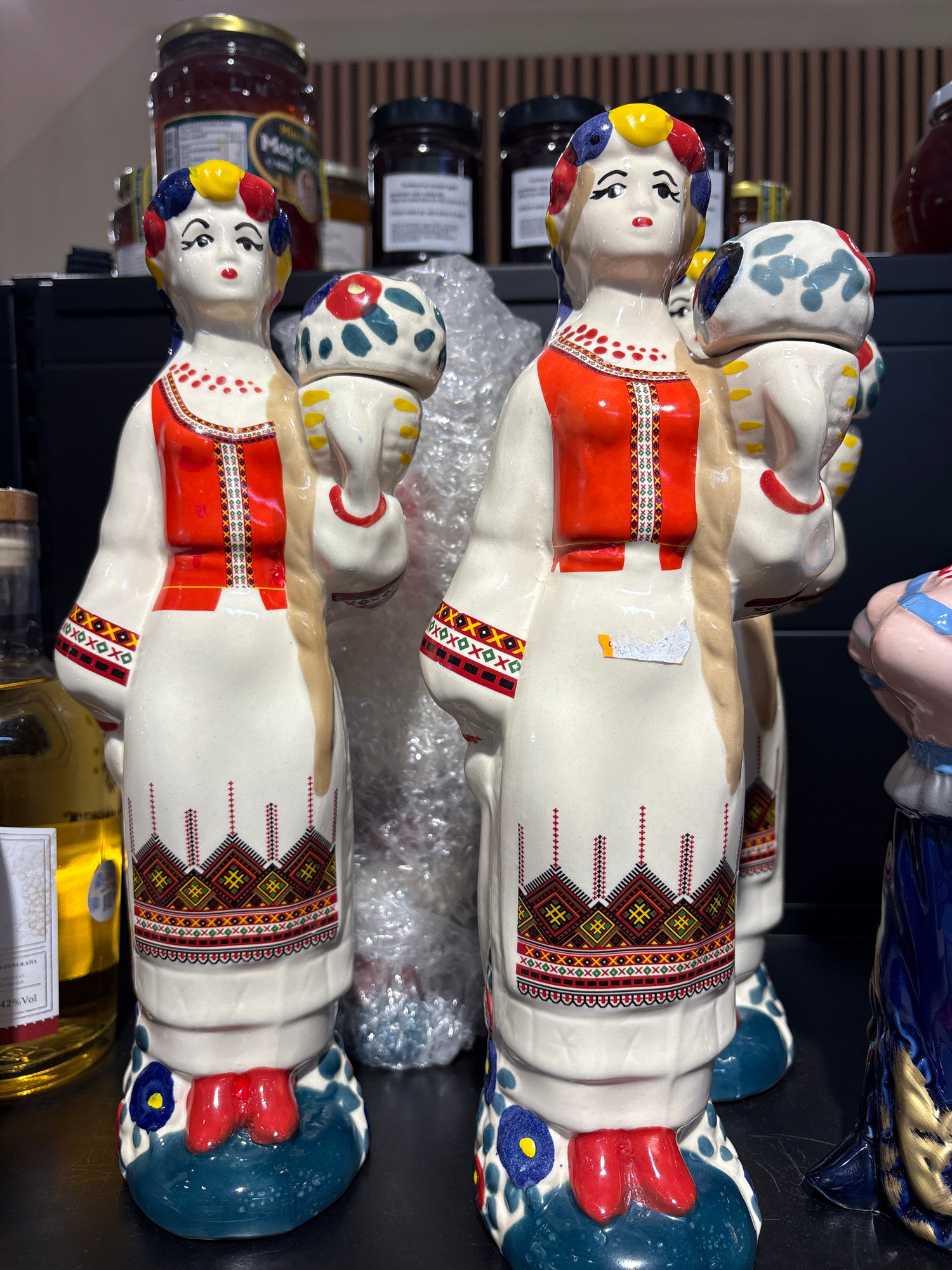 The image shows two decorative ceramic figures, likely nesting dolls, arranged vertically. The figures are painted in vibrant colors, primarily white, red, blue, yellow, and black, with intricate geometric patterns adorning their clothing. They appear to be stylized representations of people, possibly in traditional attire.
The setting seems to be a retail environment, possibly a shop or market stall, given the presence of jars on shelves in the background. The jars are of various sizes and labels, one of which is partially legible and reads "Moy Co." Another visible text, possibly from a label on one of the figures or an item nearby, includes "42% Vol" and what appears to be "XOXOXOXOX" repeated in a pattern.
The lighting suggests an indoor setting, likely during daylight hours, but no specific time of day or weather conditions can be determined. The figures themselves are the primary focus, with details like their facial features, painted clothing patterns, and headdresses clearly visible. One of the figures has a small white sticker with some orange on it, partially obscuring the pattern on its chest. The figures are placed on a dark surface, and some are wrapped in bubble wrap, suggesting they are either for sale or have been recently handled. The overall impression is one of decorative folk art or collectible items. The location context of Valencia, Spain, does not provide any specific visual cues within the image itself.