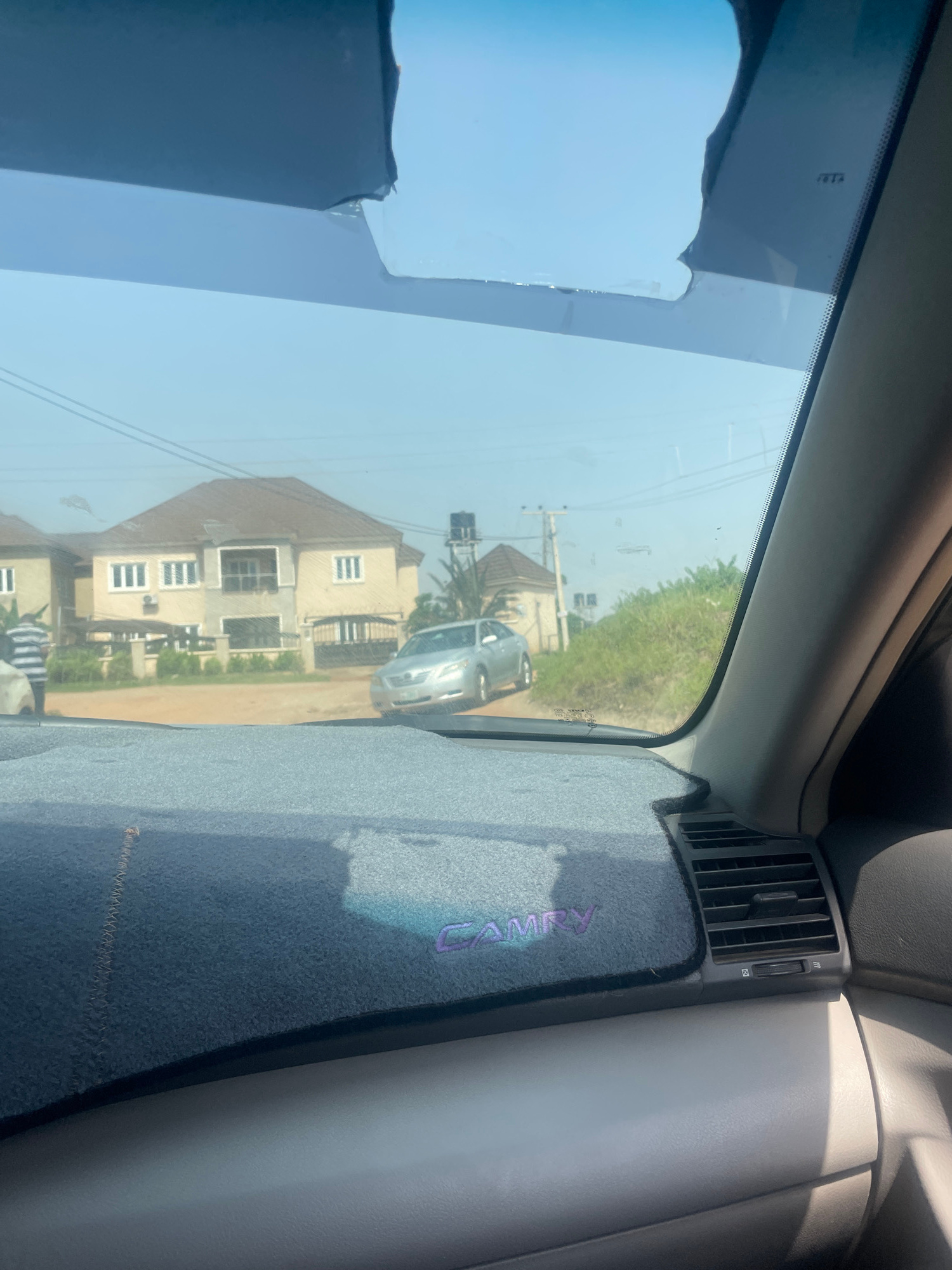 The image shows a view from inside a car, looking out through the windshield. The windshield has visible damage at the top. Outside the car, in the background, are houses, electric poles, and vegetation. A silver sedan is parked on the right side of the road. The foreground shows the car's dashboard, which is covered with a dark grey mat. The word "CAMRY" is embroidered in purple on the mat. The image appears to have been taken on a sunny day. The location is identified as Dutsen Alhaji, Nigeria.