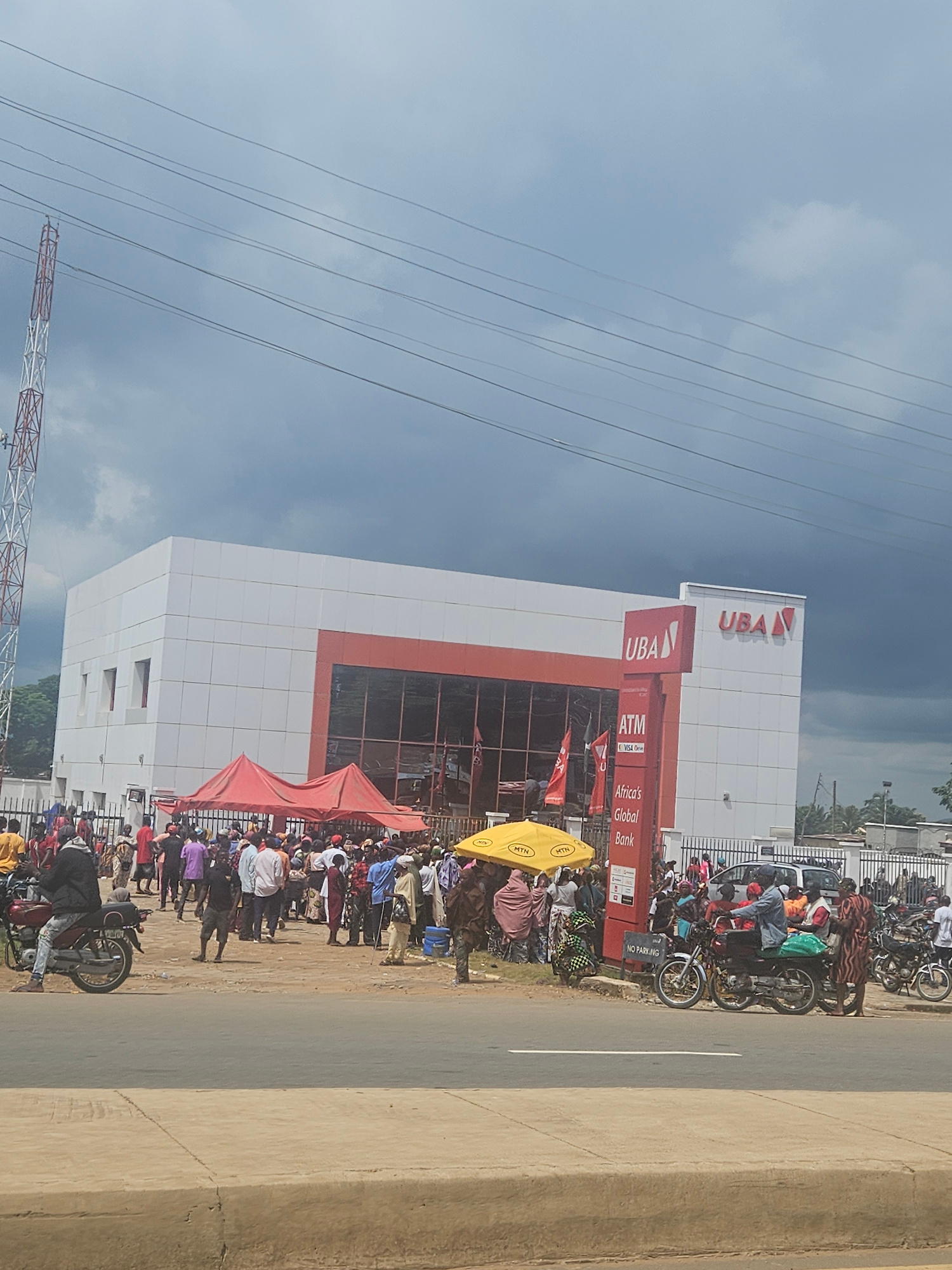 A crowd of people are gathered outside a UBA bank in Ube, Nigeria, under a cloudy sky. The bank building is modern, with a white facade and orange accents. A red canopy is set up outside the entrance, and there are several ATM machines visible. People are standing and talking, and some are on motorcycles. The scene appears to be busy, with a lot of activity.