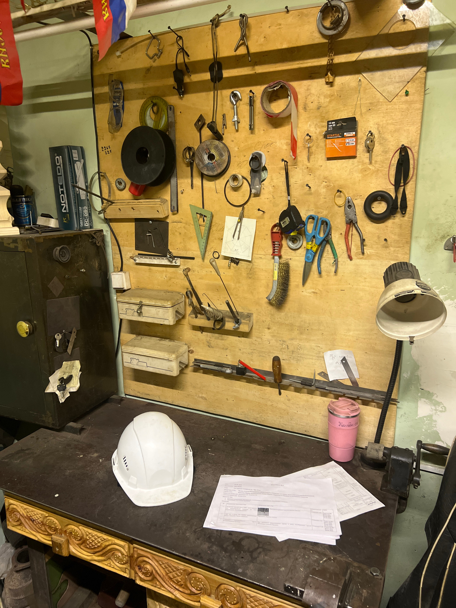 The image shows a cluttered workbench area in what appears to be a workshop or storage space. The workbench is covered with various tools, including a white hard hat, papers, and a pink thermos. Behind the workbench is a wooden board mounted on the wall, covered with an assortment of tools such as pliers, scissors, brushes, keys, and measuring devices. To the left of the workbench is a dark metal safe with keys and papers attached to it. The overall environment suggests a functional, though somewhat disorganized, workspace. There is no visible indication of the city of Nizhny Tagil, Russia, within the image.