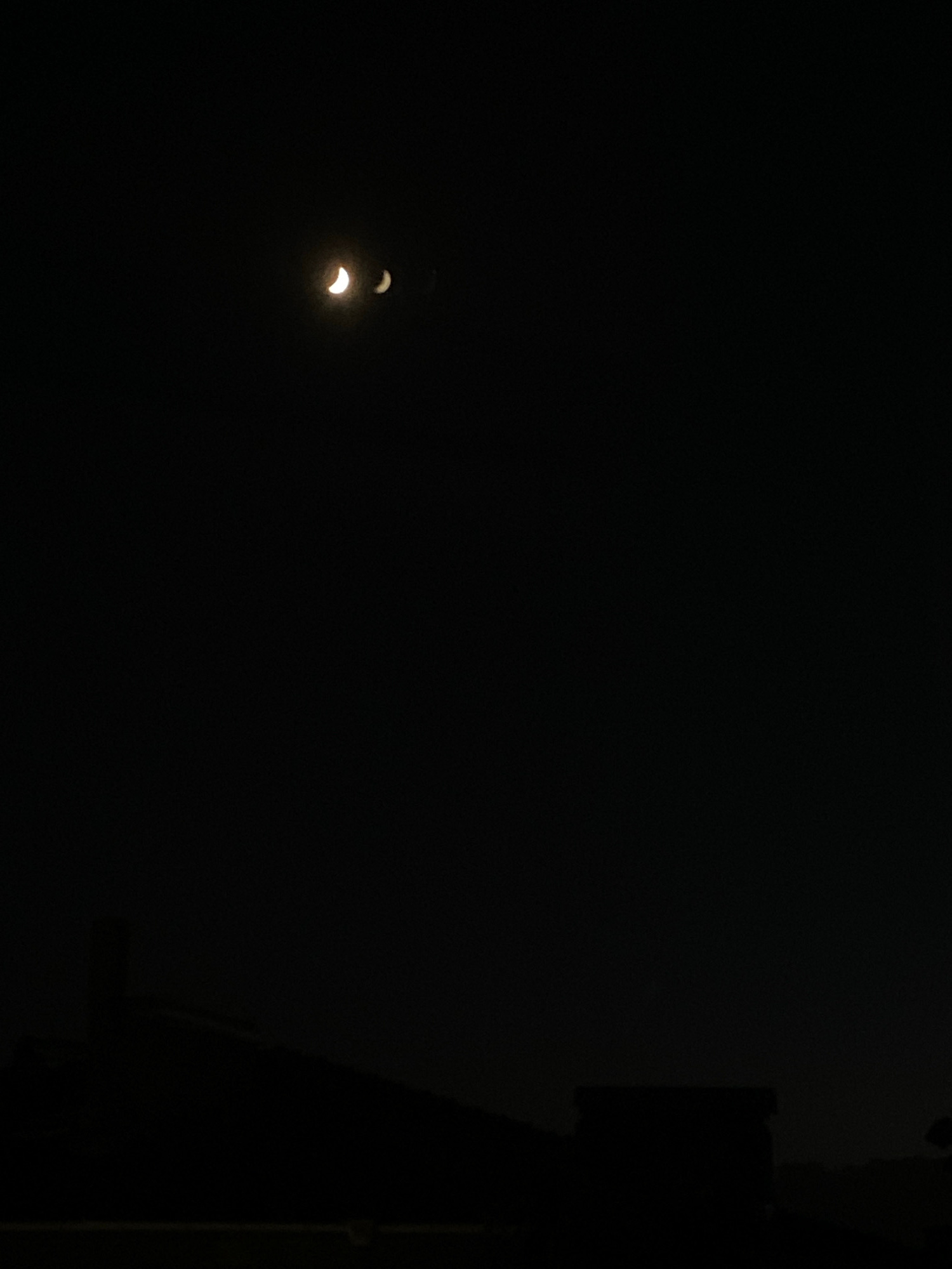 The image shows a dark night sky with two crescent moons visible. The larger moon is on the left, and the smaller moon is to its right. The moons are illuminated by an unseen light source, likely the sun, and appear as thin slivers of light against the blackness of space. Below the moons, silhouetted against the faint moonlight, is the outline of a city skyline. Buildings of various heights and shapes are visible, creating a jagged horizon. The lower portion of the image is dominated by the dark silhouettes of the city's architecture, with some structures appearing taller than others. The overall impression is one of a quiet, nocturnal urban landscape illuminated by celestial bodies.