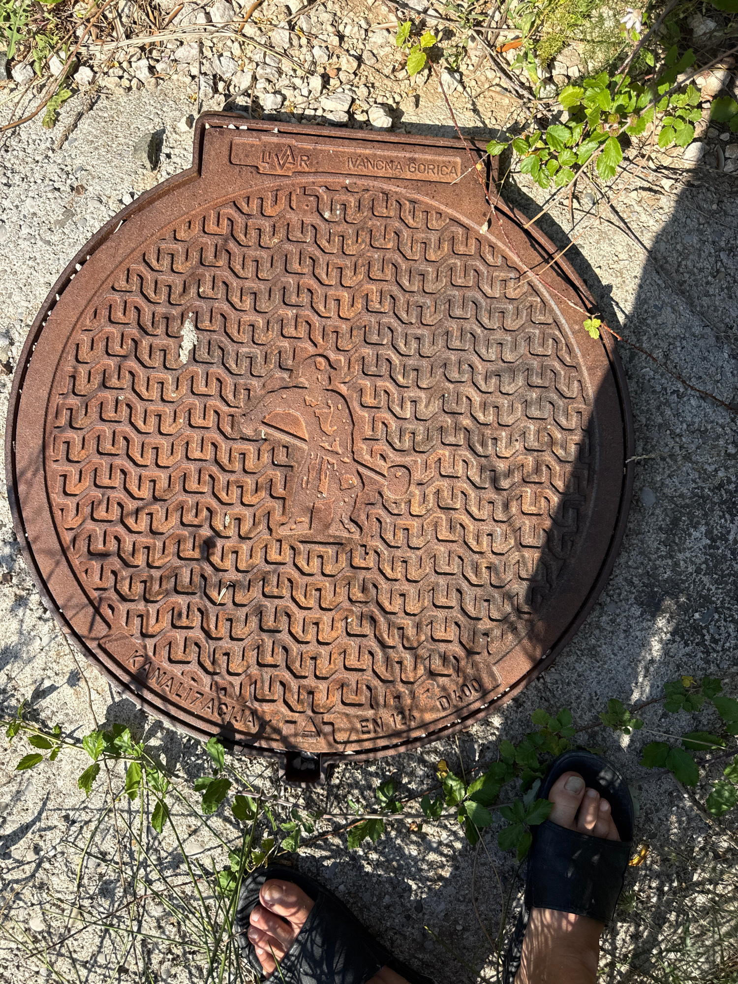 The image shows a rusty, circular manhole cover with a patterned texture and a central emblem that appears to be an animal, possibly a bird or a mythical creature. Text is embossed around the edge of the cover. The manhole cover is situated on a concrete surface with gravel and dry vegetation surrounding it. The lower right portion of the image includes a person's foot wearing a black sandal, partially obscured by shadows. The scene is outdoors, likely in an urban or semi-urban environment.
