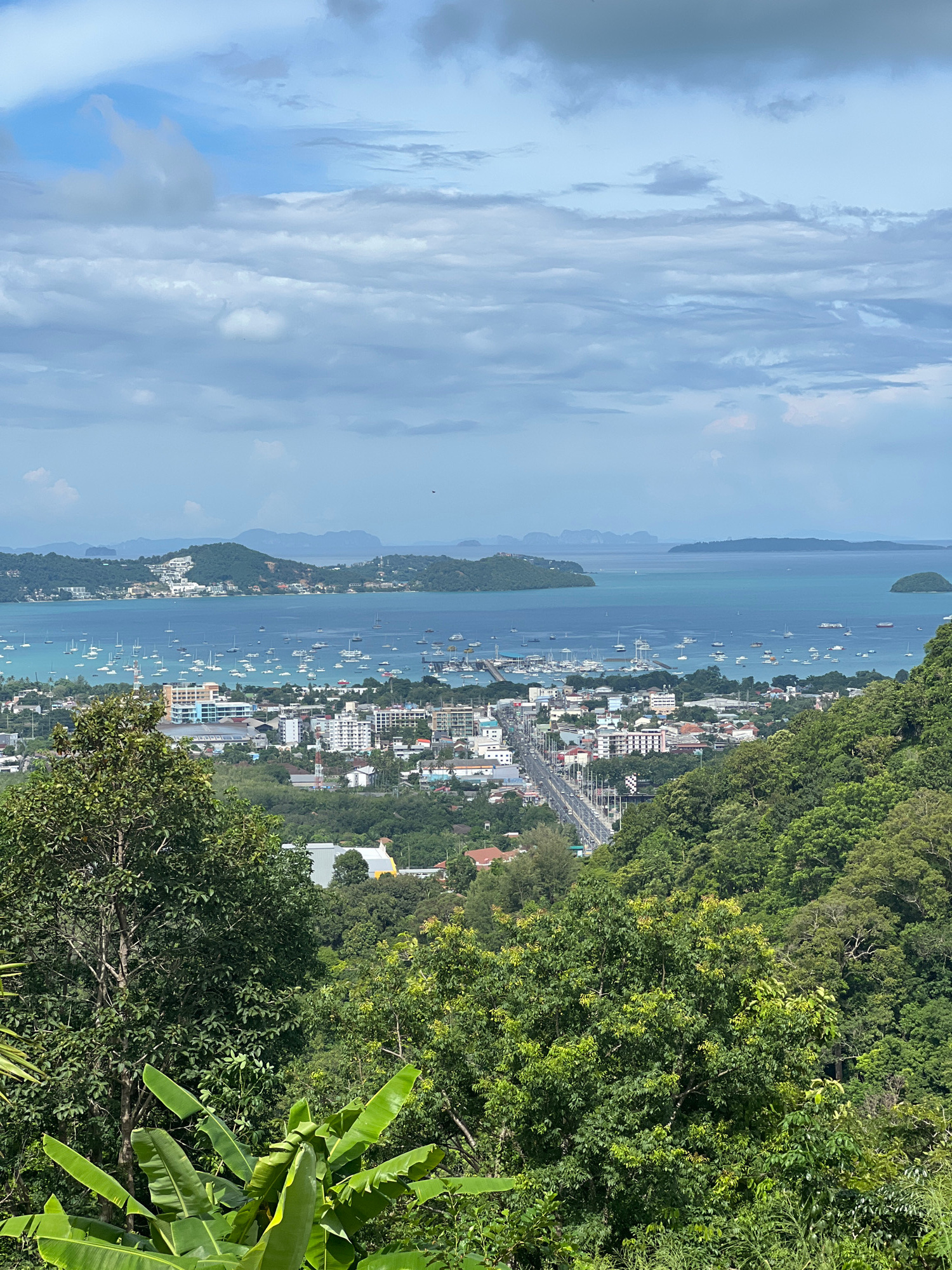 A wide-angle, elevated view captures the Chalong Bay in Chalong, Thailand, under a partly cloudy sky. The foreground is dominated by lush green trees and large banana leaves, obscuring a portion of the lower left quadrant. Beyond the foliage, a coastal town unfolds, characterized by a dense collection of buildings with white, beige, and red exteriors. A prominent multi-lane road runs through the town, marked by vehicles and streetlights.
The bay itself is a hub of activity, with numerous sailboats and yachts anchored and sailing across its turquoise waters. In the mid-ground, a modern bridge spans a section of the bay, connecting landmasses. The background reveals a series of verdant, mountainous islands scattered across the horizon, partially shrouded in a haze. The water transitions from a bright, clear turquoise near the shore to a deeper blue further out. The overall scene depicts a bustling coastal area with a mix of urban development and natural island scenery.