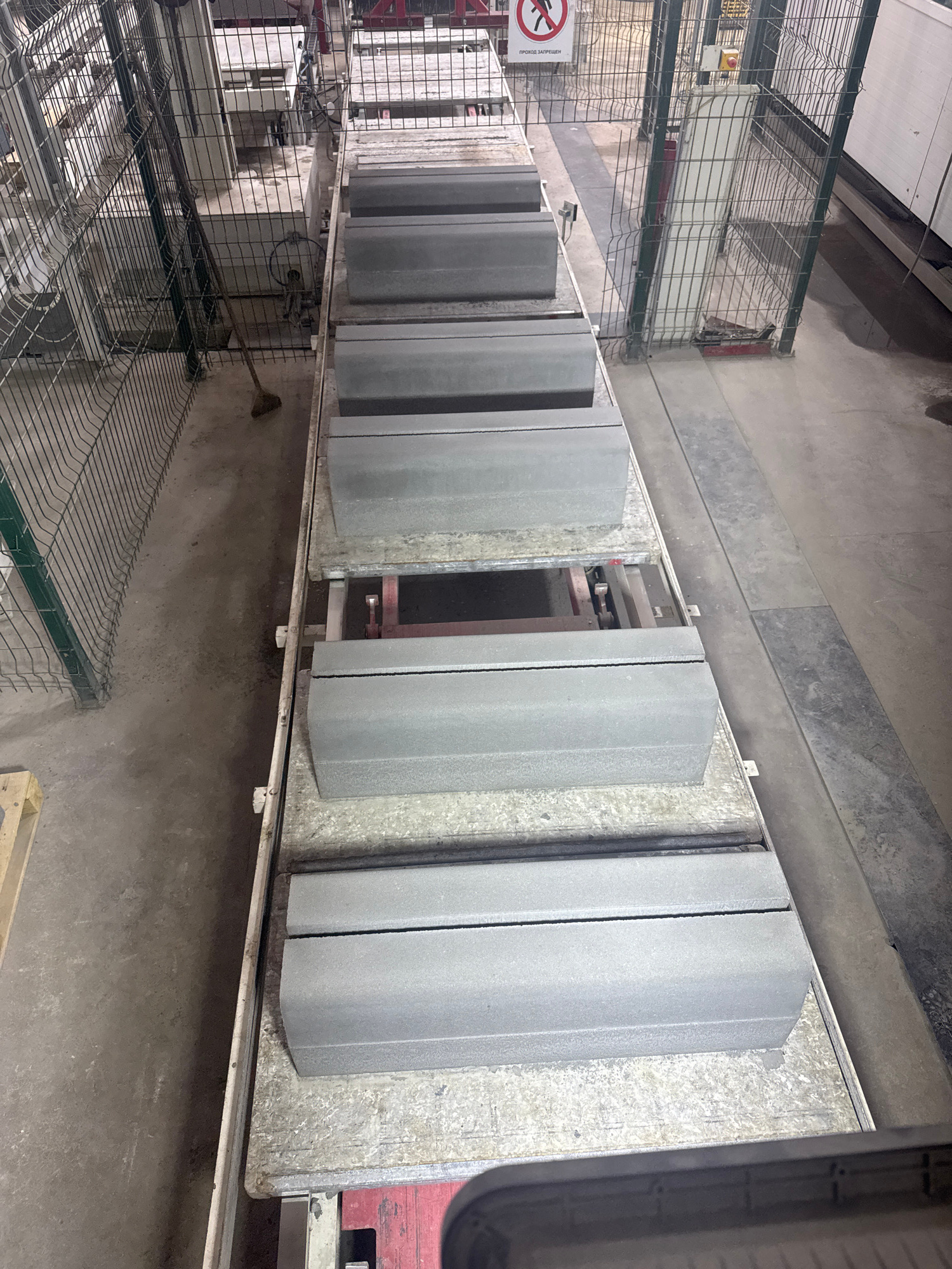 The image captures an industrial setting, likely a manufacturing facility for concrete products, in Shamalgan, Kazakhstan. A conveyor belt system is visible, carrying rows of pre-cast concrete curb stones. These stones appear uniformly grey and have a textured finish. They are arranged in multiple tiers on the conveyor, suggesting a production line in operation or awaiting processing. The environment is industrial, with concrete flooring and metal fencing present. In the background, there are glimpses of machinery and other industrial equipment. A broom lies on the floor near the fence, indicating recent or ongoing cleaning activities. A sign with red lettering and a prohibition symbol is partially visible in the upper background. The overall scene is utilitarian and focused on the production of building materials.