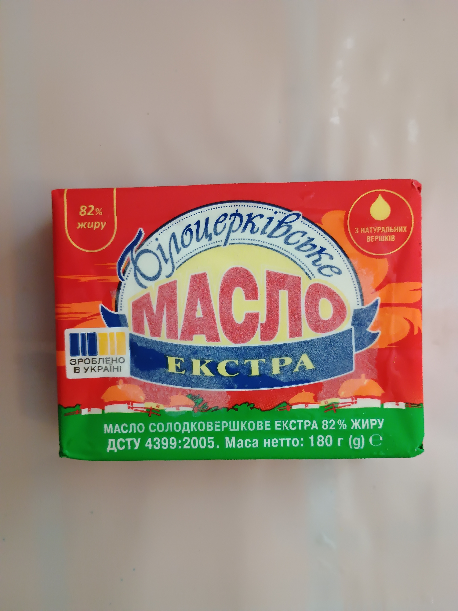 A product package for butter is displayed on a neutral, light-colored surface. The packaging is predominantly red and green, featuring Ukrainian text and graphics. The front of the package shows the brand name "Масло" (Maslo), which means "butter," in large, red, stylized letters within a yellow oval. Above this, a blue, dotted, cursive font spells out "ВолЬЦЕРКОВЬСКО" (Vol'tserkov'sko), the brand name. To the left of the brand name, "82% жиру" (82% fat) is printed in white text on a red background. Below the brand name, "ЕКСТРА" (Extra) is written in blue capital letters. A small emblem with a yellow drop signifies "3 НАТУРАЛЬНИХ ВЕРШКІВ" (From natural cream) in a circular gold outline on the upper right side. On the lower left, a blue rectangle with vertical lines indicates "ЗРОБЛЕНО В УКРАЇНІ" (Made in Ukraine). The bottom of the package is green with white text that reads "МАСЛО СОЛОДКОВЕРШКОВЕ ЕКСТРА 82% ЖИРУ" (Sweet cream butter Extra 82% fat) and "ДСТУ 4399:2005. Маса нетто: 180 г (g)" (DSTU 4399:2005. Net weight: 180 g). The image does not provide any information or context that would allow for its location to be determined in Zaporizhzhia, Ukraine, or any other city.