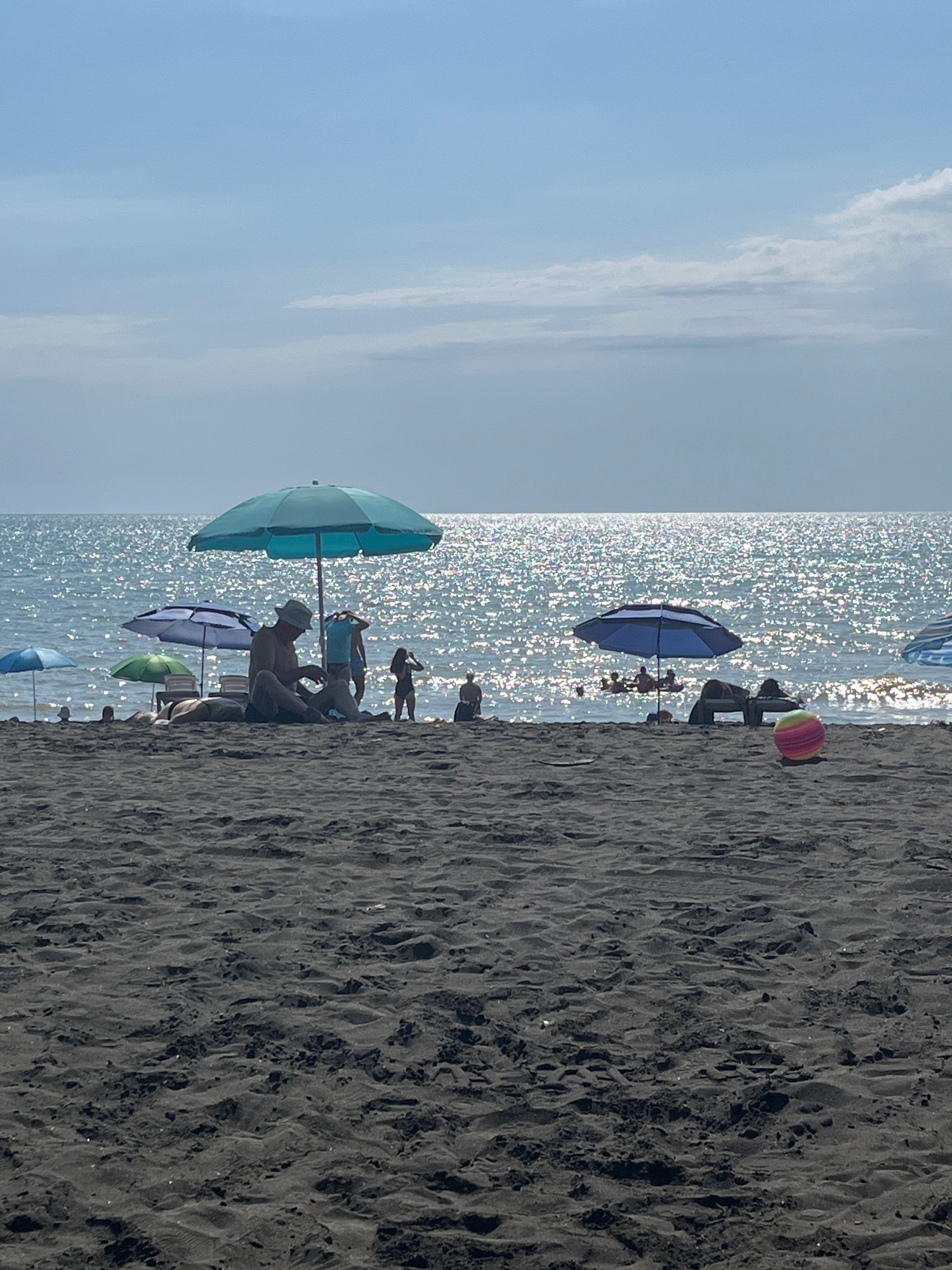 The image depicts a sunny day at Ureki beach in Georgia, characterized by dark sand in the foreground. Several beach umbrellas are set up, with people relaxing and swimming in the shimmering sea in the background. A man wearing a hat sits on the sand under a large turquoise umbrella. Other umbrellas are scattered across the beach, some with people underneath. In the water, a group of people are visible, some swimming and others on inflatable devices. A colorful striped ball rests on the sand near the water's edge. The sky is a pale blue with some scattered clouds.