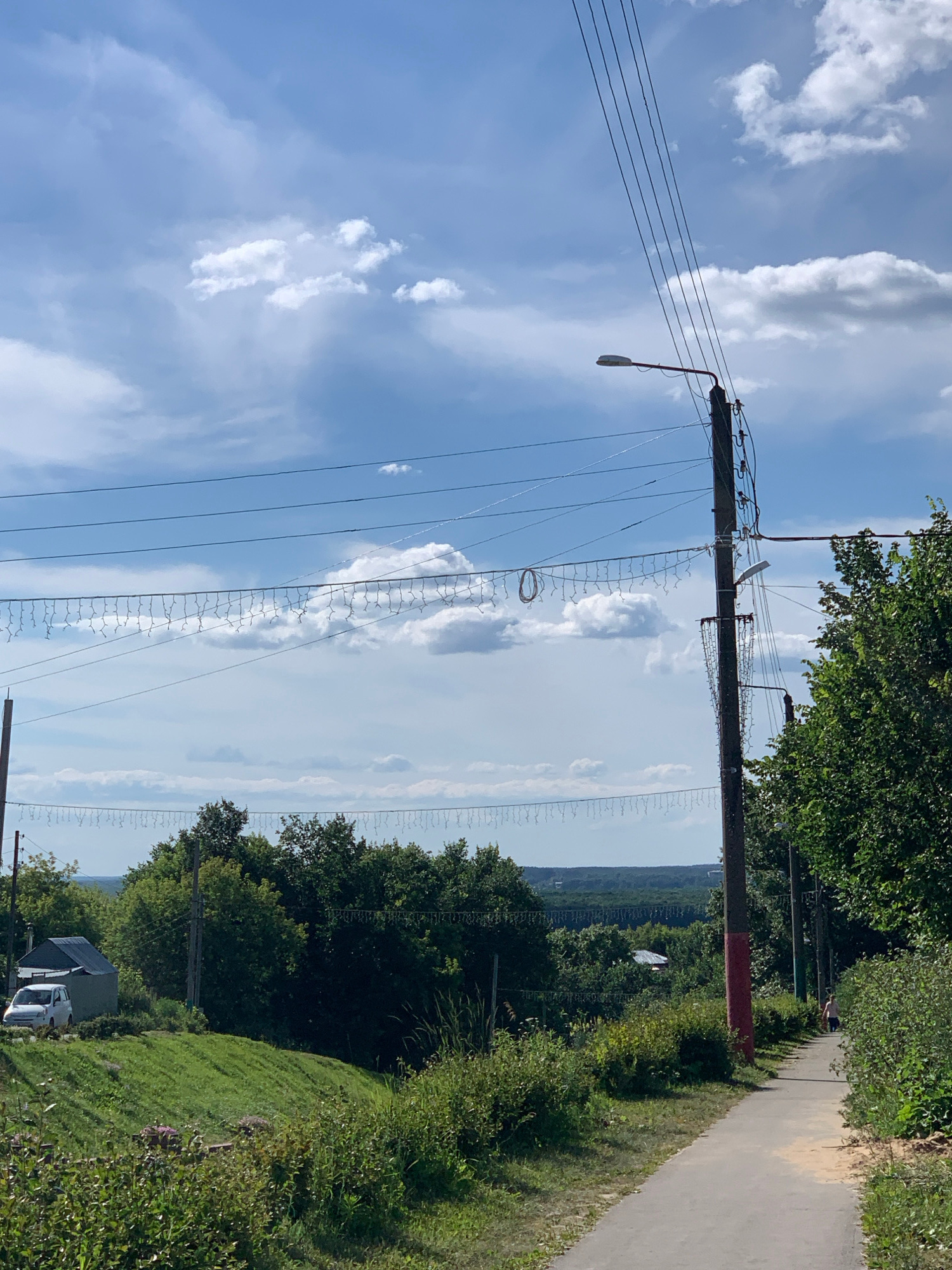 This is a medium-shot, outdoor photograph taken on a sunny day with scattered clouds in the sky. The scene appears to be a rural or suburban area on the outskirts of Pavlovo, Russia, likely during summer.
In the foreground, a paved pathway runs diagonally from the lower right to the mid-left of the frame. The pathway is bordered on the right by dense green bushes and trees. To the left of the pathway, a grassy slope rises and is covered with various plants. A white van is parked on the left side of the slope, near a small building with a dark roof.
Dominating the mid-ground and background is a dense expanse of green trees, indicating a wooded or heavily forested area. In the distance, the landscape appears to undulate, suggesting rolling hills or a valley.
Several utility poles with overhead wires are visible throughout the image, including one prominent pole in the mid-right with a street lamp attached and a red-painted section near its base. These poles support numerous electrical wires, with a string of decorative lights draped between some of them.
A lone figure, appearing to be a person walking, is visible on the pathway in the distance towards the right side of the image. The lighting suggests it is daytime, with the sun casting shadows. The overall impression is of a tranquil, natural setting with signs of human infrastructure.