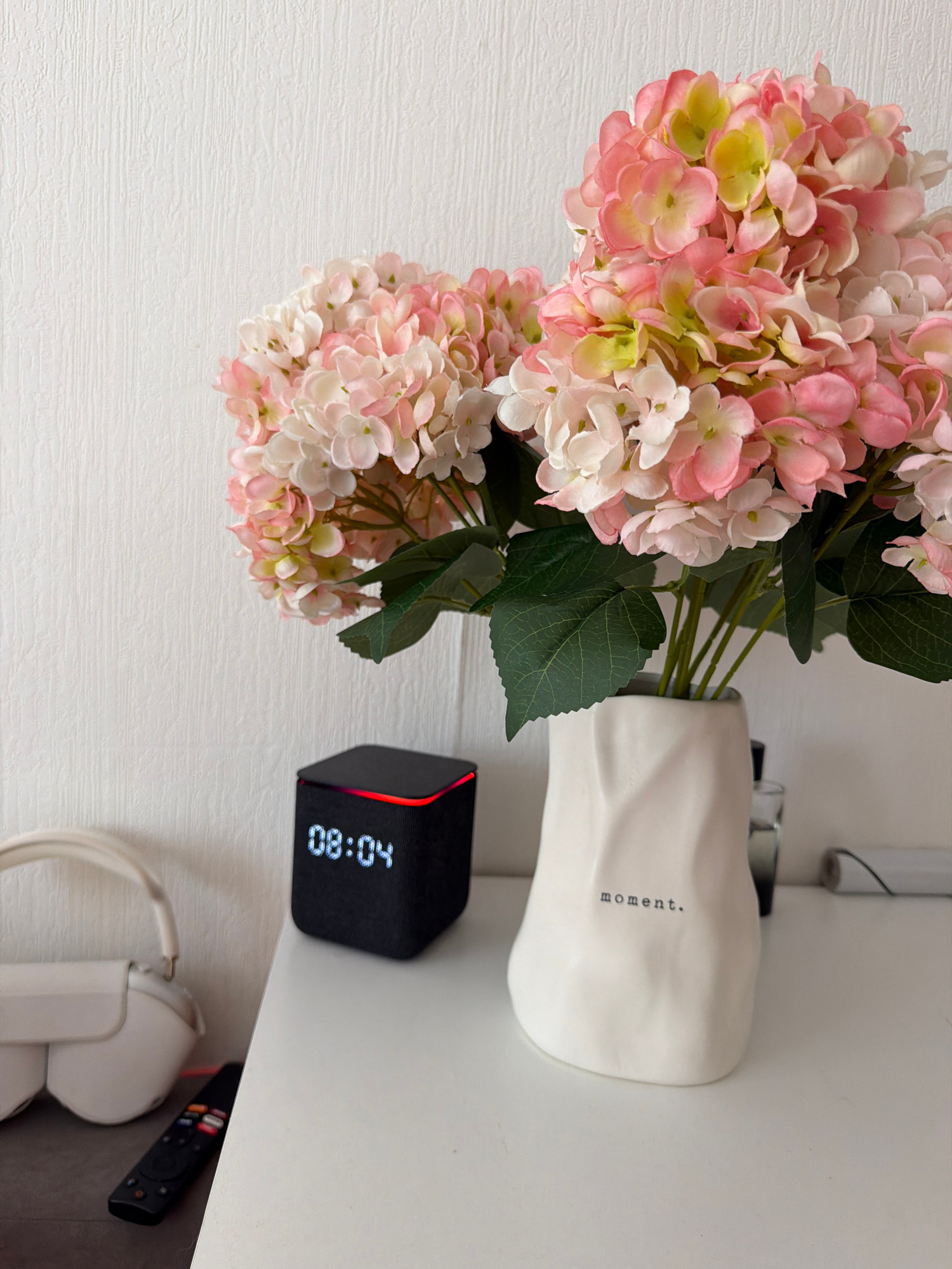 The image shows a bedside table with several objects. In the foreground, there is a vase with pink and white hydrangeas. The vase has the word "moment." printed on it. To the left of the vase, there is a black digital clock displaying "08:04". Further to the left, there is a pair of white headphones and a remote control. The background is a white, textured wall. The scene appears to be indoors, likely in a bedroom, and there is no indication of its location in Moscow, Russia.