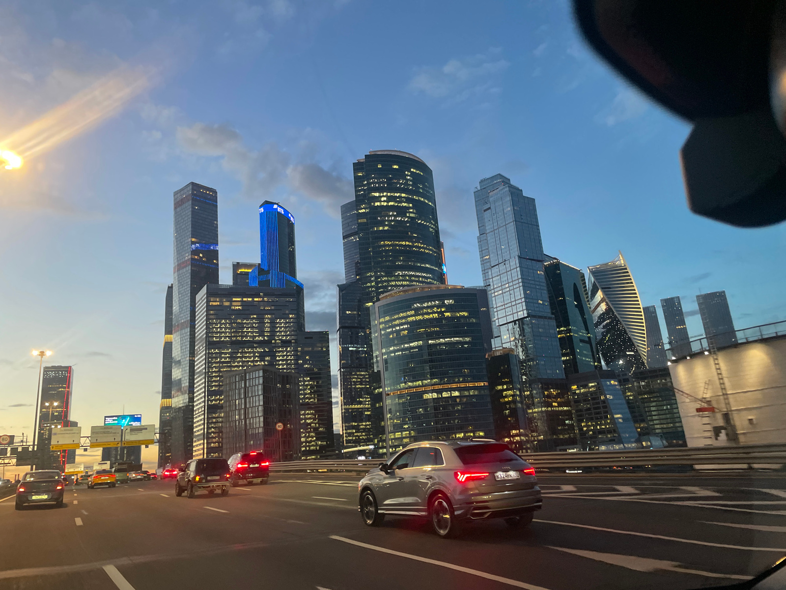The image captures a view from a car on a highway in Moscow, Russia, with the Moscow City skyline dominating the background. The sky is a twilight blue with some clouds, illuminated by the setting sun on the left. Several modern skyscrapers, many with illuminated windows, rise majestically. One building on the left is lit with bright blue vertical lights and displays the "ВТБ" logo.
On the highway, several cars are visible, moving in the same direction. A silver Audi SUV is in the foreground, moving to the right. Other vehicles, including sedans and SUVs, are further ahead in traffic. Traffic signs, including speed limit signs (80), are present along the roadside. The highway is multi-lane, with clear lane markings. The overall scene depicts an urban environment at dusk, with city lights beginning to illuminate the buildings and vehicles navigating the busy highway.