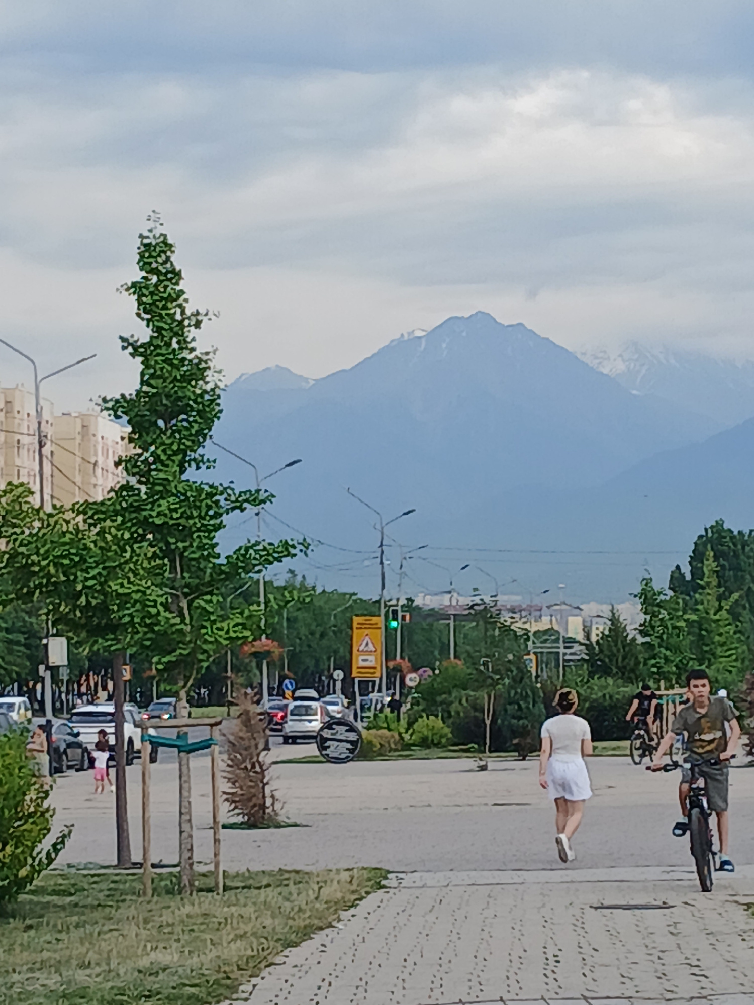 A vertically oriented, medium-shot photograph captures a street scene with a mountain range in the background. The sky is overcast with diffused light.
In the foreground, a paved sidewalk runs along a street where several cars are visible, some parked and others in motion. A woman in a white dress is walking on the sidewalk, her back to the camera. Further down the sidewalk, two people are on bicycles, one a man in a green shirt and shorts, the other a person whose gender is unclear, also on a bike.
Green trees line the street, with foliage obscuring parts of the buildings visible in the distance. The buildings appear to be of medium height and are light-colored. Traffic lights are visible, with one showing green. There are several streetlights and poles along the street.
A large, mist-covered mountain range dominates the background, its peaks appearing snow-capped. The overall impression is of a casual day in an urban environment with a natural backdrop. The time of day appears to be daytime, given the ambient light. There is no discernible text in the image.