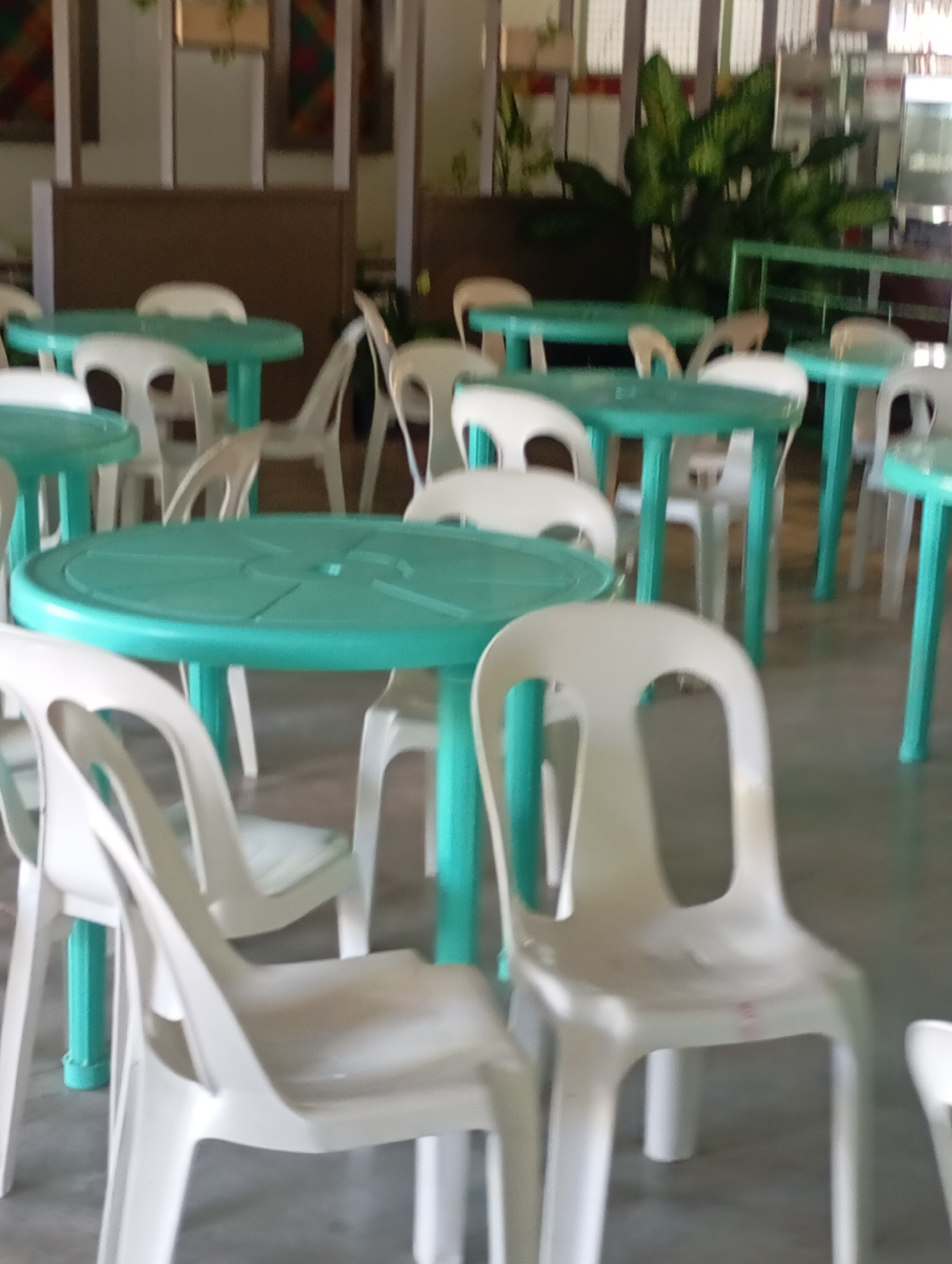 The image shows an indoor seating area with multiple round, teal-colored tables and white plastic chairs. The tables have a slightly raised edge and appear to be made of plastic. The chairs are standard stackable plastic chairs with curved backs and seats. Some chairs are tucked under the tables, while others are pulled out. In the background, there are hints of a wall with decorative panels and large green plants. The overall impression is of a casual dining or waiting area, possibly in a restaurant or food court. The lighting is diffused, suggesting an indoor environment. Given the style of furniture and the likely climate, this scene is consistent with establishments found in Davao City, Philippines.
