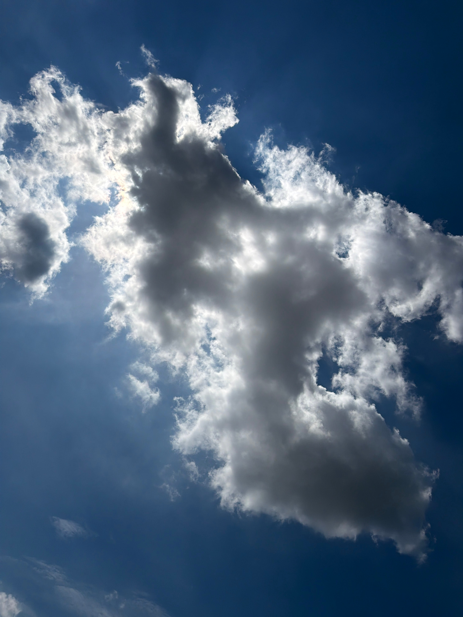 A close-up, vertical shot captures a dramatic sky with dark, heavy clouds and bright, sunlit clouds against a deep blue background. The sun is partially obscured by a large, dense cloud in the center, casting rays of light through the thinner parts of the cloud formations. The overall impression is one of dynamic weather. The image is located in Męcina, Poland. There are no people or specific objects visible, only the natural environment of the sky and clouds.