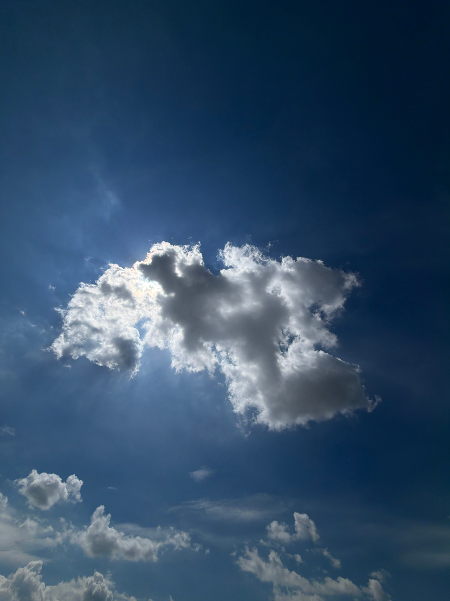 The image captures a bright blue sky filled with scattered clouds. A large, dense cloud formation dominates the center, partially obscuring the sun, which creates a bright halo and visible sunbeams radiating outwards. Smaller, wispy clouds are visible in the lower portion of the frame. The overall scene depicts a sunny day with a dynamic sky. The location in Męcina, Poland, is not visually represented in the image.