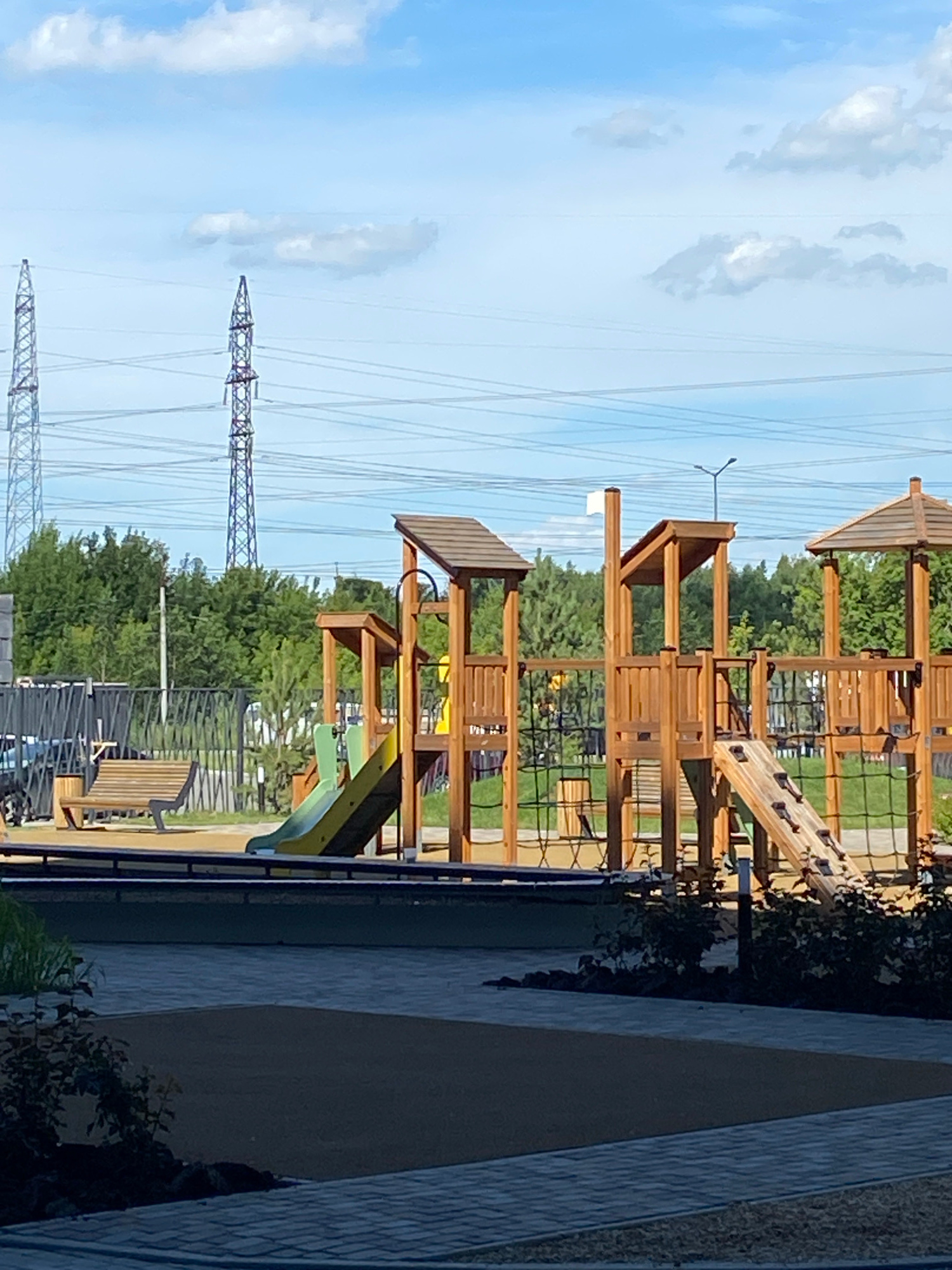 This image shows a modern wooden playground with multiple slides, climbing structures, and a bench. The playground is situated in an outdoor urban environment, with a fence bordering one side and lush green trees in the background. In the distance, tall electrical towers and power lines are visible against a bright blue sky with scattered clouds. The foreground features paved pathways and landscaping. No people are present in the image. The scene appears to be in Chelyabinsk, Russia.
