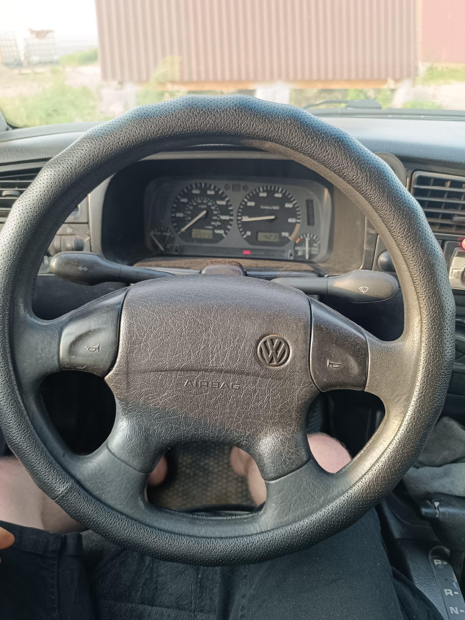 The image shows a close-up view of a car's interior, focusing on the steering wheel and dashboard. A person's legs, wearing black pants, are visible below the steering wheel, indicating they are in the driver's seat. The steering wheel is covered in a textured black material and has the Volkswagen logo in the center. The dashboard features a speedometer, tachometer, and other gauges. A windshield wiper control stalk is visible to the left of the steering wheel. Outside the car, a building with corrugated siding and some greenery can be seen through the windshield. The lighting suggests it is daytime, possibly late afternoon, and the weather appears to be clear. The scene appears to be in a rural or semi-rural area, consistent with the location context of Qonaev, Kazakhstan. There is no specific activity or event depicted other than the implied act of driving or preparing to drive. The visible text includes "AIRBAG" on the steering wheel, the Volkswagen logo, and markings on the gear shift.