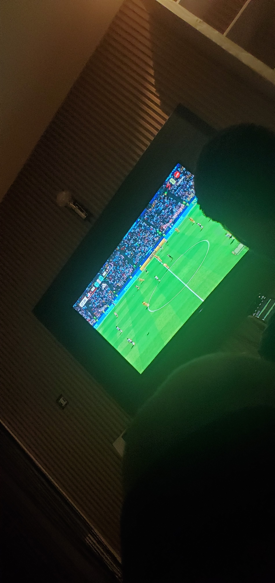 An angled, medium shot captures a television screen displaying a soccer match. The screen is illuminated and shows a green field with white markings and numerous figures representing players and spectators. A person's dark silhouette is visible in the lower right corner, obscuring a portion of the television and the immediate foreground. The background consists of a textured, ribbed wall. The setting appears to be indoors, likely a living room or entertainment space. There is no identifiable information to place this scene in Gura Top, Nigeria.