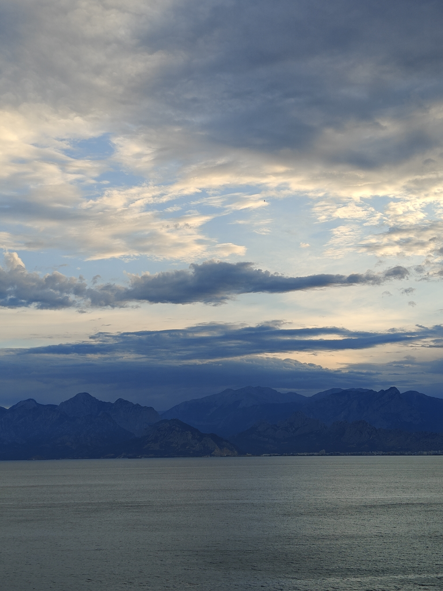 The image captures a serene seascape with a dramatic sky and a mountain range in the background. The sea occupies the lower half of the frame, its surface textured with gentle ripples that reflect the muted light. In the distance, a chain of dark, jagged mountains stretches across the horizon, their peaks softened by the atmospheric conditions. Above the mountains, a vast sky is filled with an array of clouds. Some clouds are light and wispy, allowing patches of pale blue sky to peek through, while others are darker and more dense, creating a sense of depth and movement. The lighting suggests it is either early morning or late evening, with soft, diffused sunlight illuminating the scene. A tiny, dark silhouette of a bird is visible in the upper portion of the sky, adding a sense of scale. The overall impression is one of tranquility and natural beauty. This scene is consistent with views of the Mediterranean coast near Antalya, Turkey, where the Taurus Mountains meet the sea, often under a sky characterized by such cloud formations.