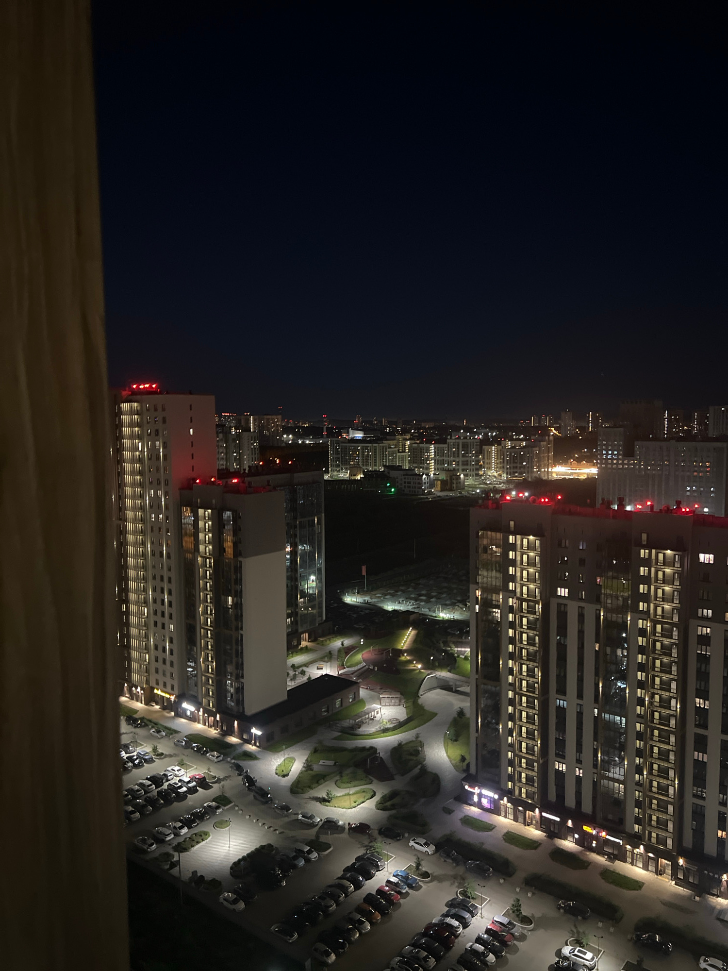 An elevated, nighttime, vertical shot captures a residential complex in Yekaterinburg, Russia, with a curtain in the foreground on the left. The buildings are tall, modern, and illuminated from within, with red lights visible on the rooftops of several structures. A street and parking lots filled with cars are visible at the base of the buildings, along with landscaped areas and walking paths. The sky is dark and clear, with no stars or moon visible. The scene conveys a sense of urban life and activity at night.